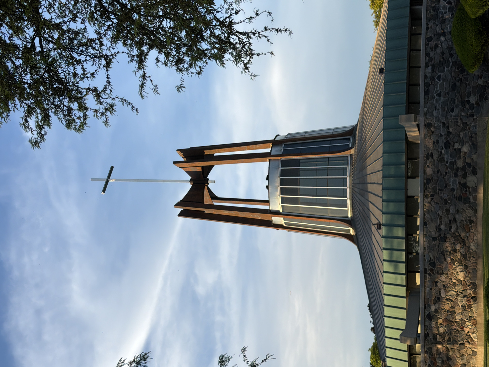 The image depicts a low-angle view of the Prince of Peace Parish church building in Niles, Illinois, USA. The building features a modern architectural style with a distinctive cross atop a brown wooden framework. The church incorporates a cylindrical structure with windows and a sloping roof with a stone exterior wall. The sky behind the building is blue with streaks of white clouds. Tree branches and foliage are partially visible in the upper and lower portions of the image.