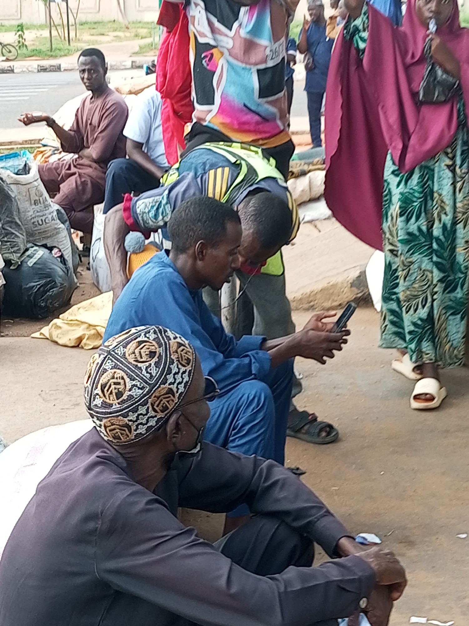 The media file shows a group of people sitting on the ground in what appears to be a market or a street setting. The subjects include several men and a woman. One man in a maroon robe is seated and looking towards the viewer with his hand outstretched, possibly begging. Another man in a blue tunic is seated and looking at a smartphone. A third man, wearing a dark outfit and an ornate cap, is also seated and appears to be looking down. A woman in a pink hijab and a green and white patterned dress is standing. Behind them, other individuals are visible, some in colorful clothing. The ground is dusty and there are various objects scattered around, including bags that look like they contain sugar. There is a bicycle visible in the background, suggesting an outdoor environment. The time of day appears to be daytime, with ambient lighting. The overall atmosphere seems casual, with people resting or engaged in their activities. There is no clear indication of a specific event, but rather a snapshot of daily life. Visible text on one of the bags reads "BLIA SUGAR". The location context is Gombe, Nigeria.
