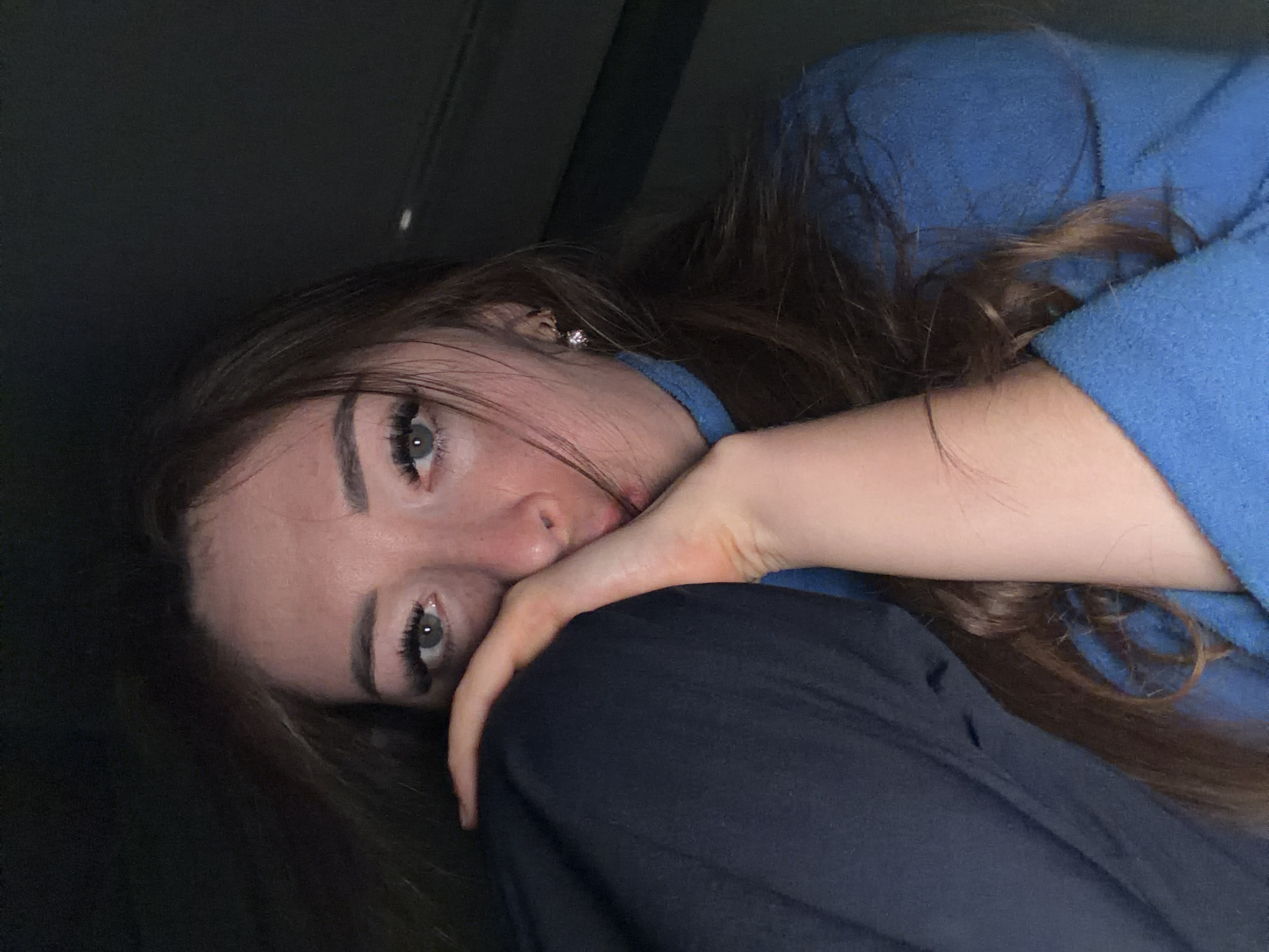 This is a close-up, low-angle, indoor photograph of a young woman. She has long, brown hair that is falling around her face. She is wearing a blue hooded sweatshirt and dark pants. Her face is partially obscured as she rests her head on her arm and knees. Her eyes are open and looking towards the camera, with long eyelashes. She is wearing a small, sparkling stud earring in her visible ear. The lighting is dim, suggesting it might be evening or a poorly lit indoor space. The woman's expression appears thoughtful or perhaps a little melancholic. The background is dark and indistinct, offering no specific clues about the location beyond the fact that it is indoors. The context provided places the image in Yarykovo, Russia. There is no visible text in the image.