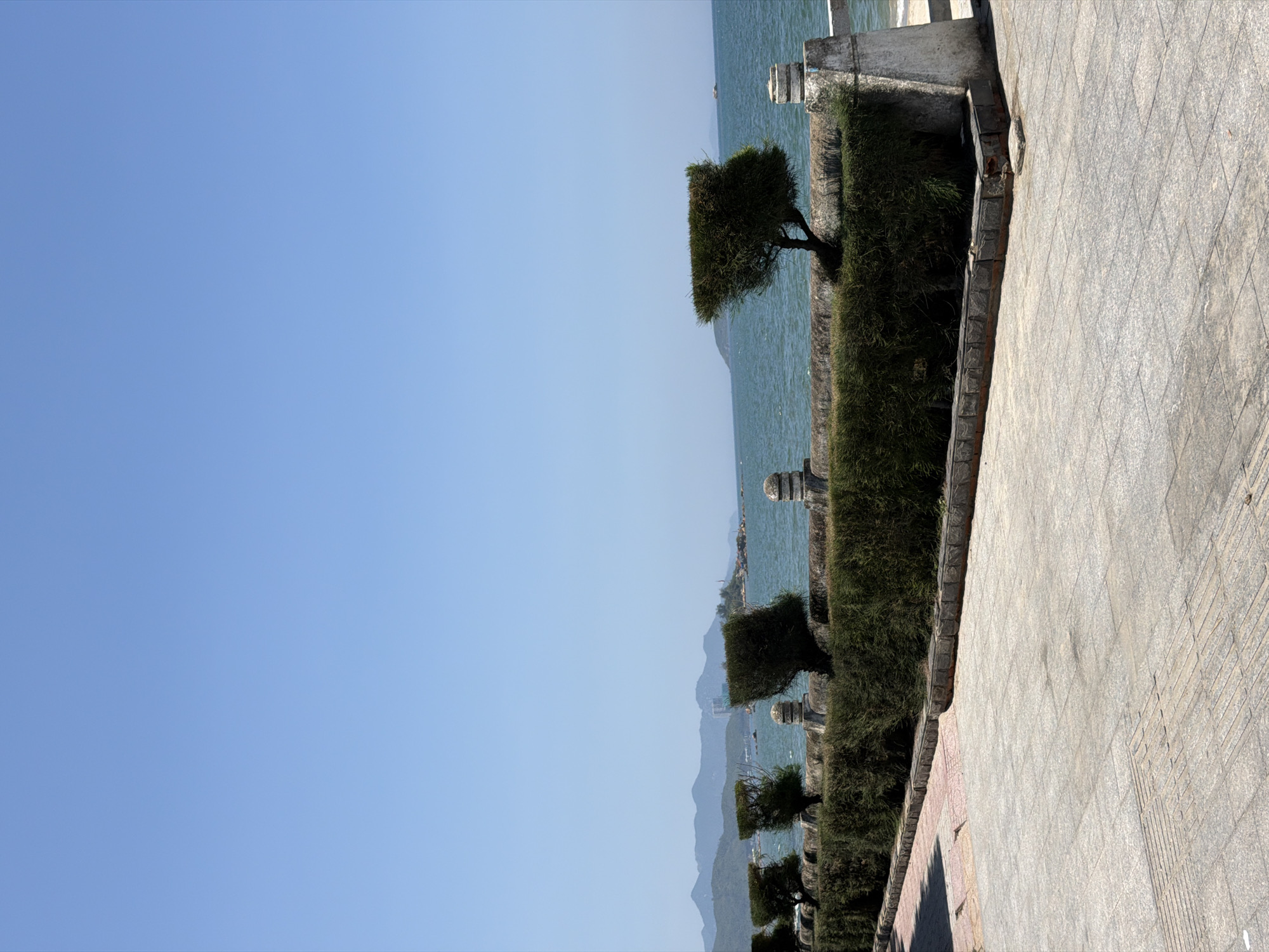 The image depicts a sunlit coastal scene in Nha Trang Ward, Vietnam, viewed from a paved promenade. The upper two-thirds of the frame are dominated by a clear, bright blue sky, fading slightly to a lighter hue near the horizon. Below the sky, the dark blue-green sea extends to a distant mountainous coastline on the left and several smaller islands or landmasses further out to the right. A tall, light-colored building structure is visible on one of the more prominent distant land features. One small, dark vessel is faintly discernible on the water towards the right horizon.
In the midground, running horizontally across the frame, is a low, robust stone or concrete barrier topped with a dense, dark green hedge. Several small, windswept or topiary-shaped trees, characterized by flattened, somewhat rectangular crowns of dark green foliage, emerge from behind or within this hedge at regular intervals. The barrier itself features several rounded, light-colored stone finials or posts spaced along its length.
The foreground consists of a grey tiled or paved walkway, composed of rectangular and square pavers. A section of tactile paving with raised parallel lines is visible in the lower left, transitioning to a raised dark stone curb that borders the hedge-topped barrier. Shadows are cast by the hedge and barrier, indicating bright, high-angle sunlight. No people, animals, or immediate human activities are present in the frame.