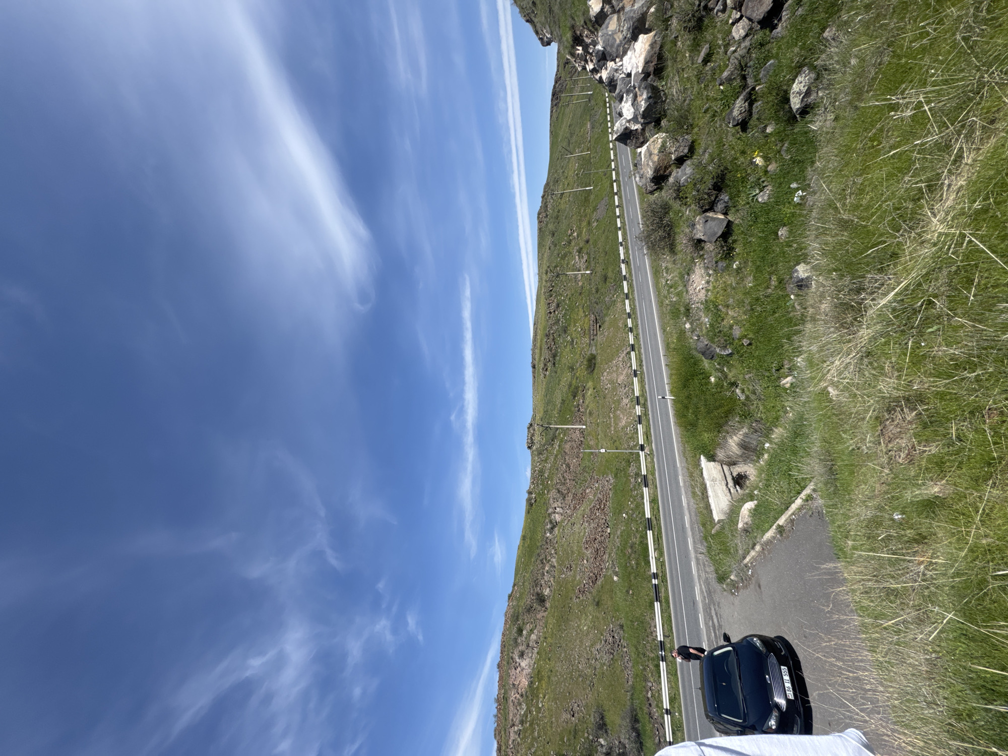A slightly rotated, eye-level shot captures a section of road near Bagravan, Armenia. To the right of the road, a steep grassy hill dotted with rocks rises, transitioning to a rocky cliff face in the upper portion. Utility poles run along the roadside, and the sky on the left side is a clear blue with streaks of clouds. A dark-colored sedan is parked near the edge of the road, and a person is standing near the car. The foreground is blurred and obscured by a light-colored object.