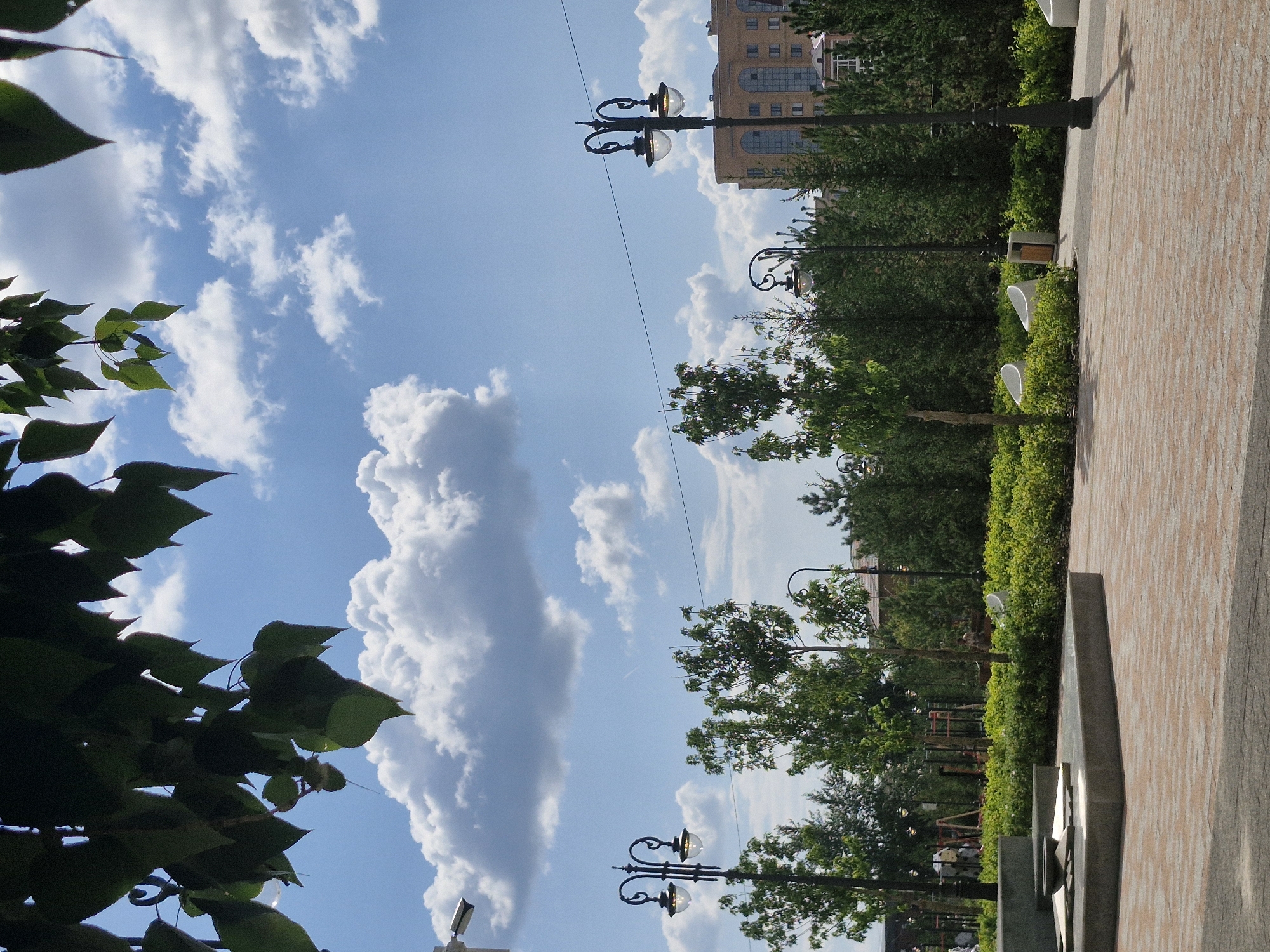 The image depicts a public park area in Kokshetau, Kazakhstan, on a bright, cloudy day. The foreground features a paved area with brick-like patterns, leading to a raised, modern monument with a bowl-shaped element on top. Behind this monument is a lush green hedge, behind which several young trees are planted. Further back, ornate lampposts with two lights each are visible, along with a line of dense evergreen trees and taller deciduous trees. In the distance, a multi-story residential building with a light-colored facade and arched windows can be seen. The sky is blue with scattered white cumulus clouds. A security camera is mounted on a pole in the upper left corner, partially obscured by dark green leaves hanging from a tree branch.