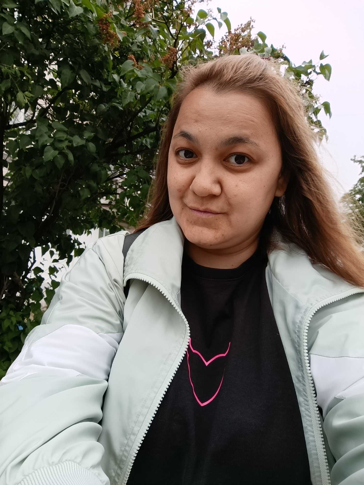 A close-up, eye-level selfie features a person, appearing to be an adult female, with long, light brown hair. She looks directly at the camera with a neutral expression, showing a slight smirk or closed-mouth smile. Her hair is slightly windswept on the right side of the frame.
She is dressed in a light mint green or pale teal zip-up jacket, which has white panels on the shoulders and inner sleeves. Underneath the jacket, a black t-shirt with a bright pink, outlined heart-like design is visible on the chest. A dark backpack strap can be seen over her left shoulder.
The background consists of dense green foliage from trees, some bearing brown clusters of what appear to be dried flowers or seeds. The sky above is overcast and bright, indicating a cloudy daytime. The setting is outdoors, likely a park, garden, or a street lined with trees, creating a natural backdrop. No text is visible in the image. The scene is casual and takes place in an outdoor environment, with the provided context suggesting it may be in Chaikovsky, Russia.