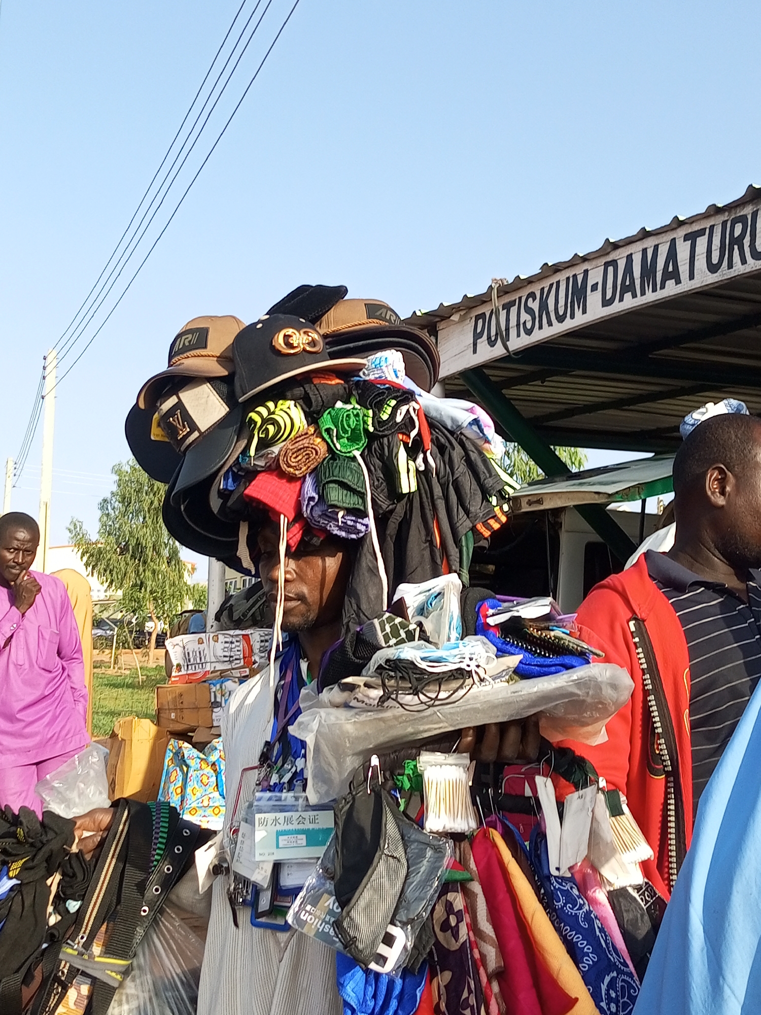 A street vendor balances an assortment of caps and folded garments on his head, his face partially obscured by the merchandise. He is standing in front of a stall with a corrugated metal roof, on which is painted "POTISKUM-DAMATURU". To his left, a man in a bright pink traditional outfit stands with his hand to his chin, looking thoughtfully at the vendor. The background shows a street scene with trees and a clear blue sky. The vendor appears to be in a market or roadside stall area.