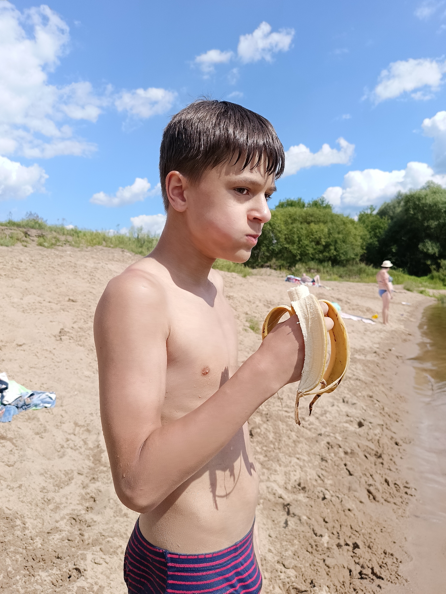 A young boy with dark hair, shirtless, stands on a sandy beach, holding a partially peeled banana. He wears striped swim trunks. His expression is thoughtful as he looks away from the camera.
The background shows a body of water, likely a river or lake, with gentle waves lapping the shore. Further back, other beachgoers are visible, including a woman in a blue swimsuit and a straw hat, and others lying on towels. Lush green trees line the opposite bank of the water. The beach itself is sandy with some vegetation on a slight incline behind it.
The sky is a bright blue with scattered white clouds, indicating a sunny day. The presence of swimsuits and the beach setting suggest it is a warm season, likely summer. The overall scene evokes a relaxed, casual atmosphere of a day spent outdoors by the water. No visible text is present in the image.