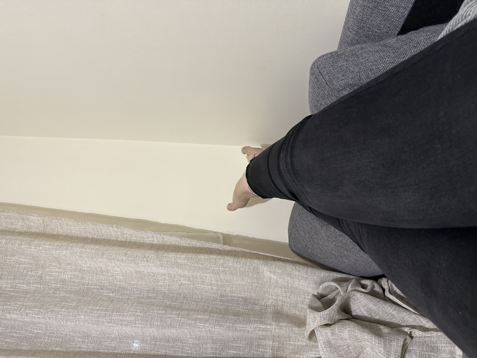 A person wearing black jeans sits on a grey upholstered chair. Their right hand extends toward a pale yellow wall. Below the wall, a light-colored, textured curtain drapes. The scene is indoors. The location is unknown, and there is no information indicating the city of Sofia, Bulgaria.