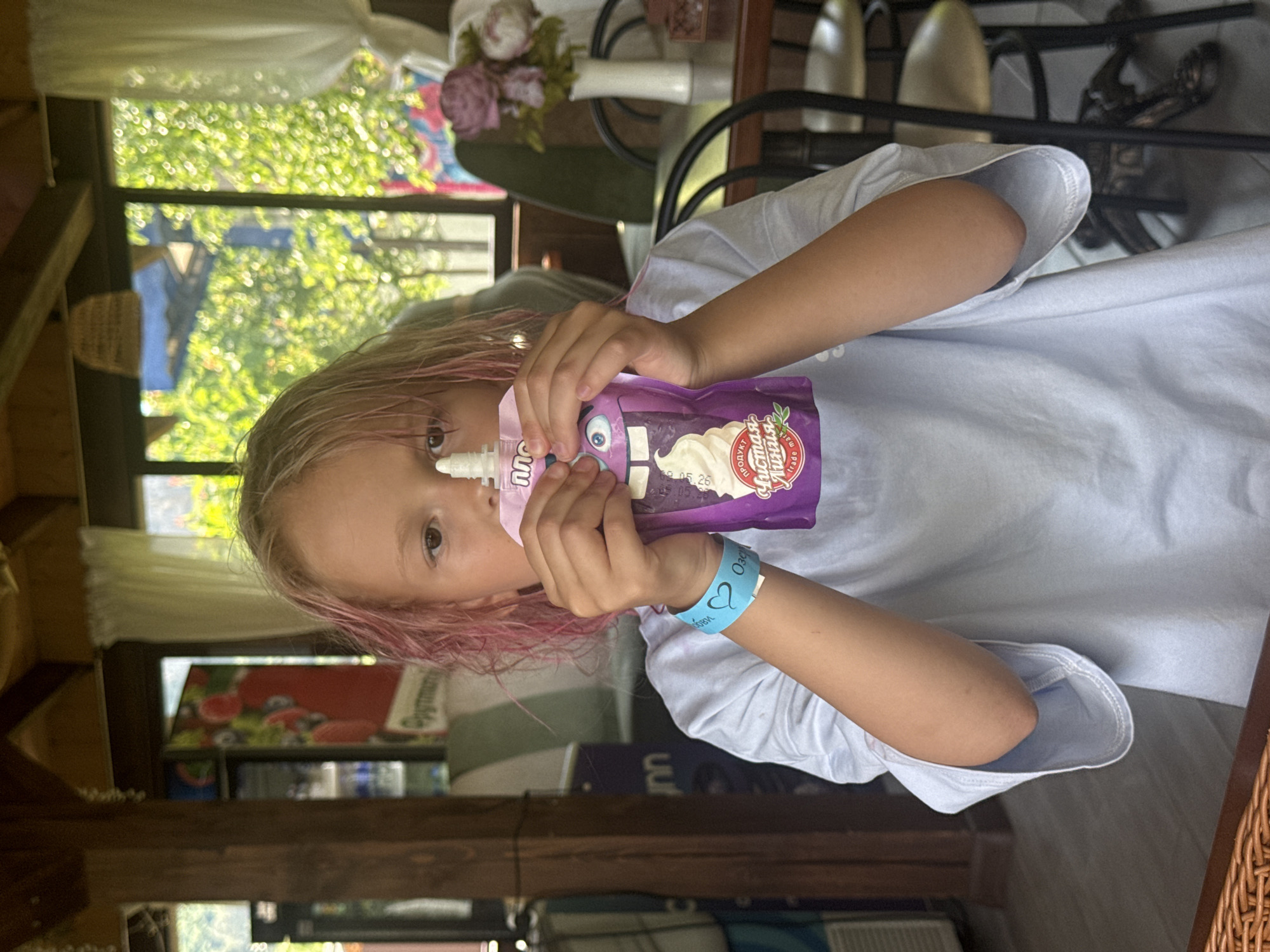 A young individual with blonde hair featuring pink dyed ends is seated indoors, looking directly forward. They are holding a purple squeeze pouch with both hands, positioned in front of their lower face. The pouch displays a cartoon character with a single large eye and sharp teeth, along with a white swirl resembling whipped cream. Cyrillic text on the pouch includes "ПРОДУКТ ЛИСТАЯ ЛИНИЯ" and "trade mark". The individual wears a light blue or grey short-sleeved t-shirt and a light blue wristband on their left wrist, featuring a white heart symbol and Cyrillic text that appears to include "ОЗОРНЫЙ".
The setting is an interior space characterized by wooden support beams and columns, suggesting a rustic or open-air structure. In the background on the left, a dark vertical panel is visible, alongside a refrigerator with colorful imagery of berries or fruit and the Cyrillic word "Орум" on its base. On the right, a table with dark metal chairs and a white vase containing artificial purple and white flowers is present. A large window with white sheer curtains overlooks an outdoor scene of dense green foliage. A light-colored woven lampshade hangs from the ceiling. The floor beneath the table appears to be a light, possibly tiled, surface.