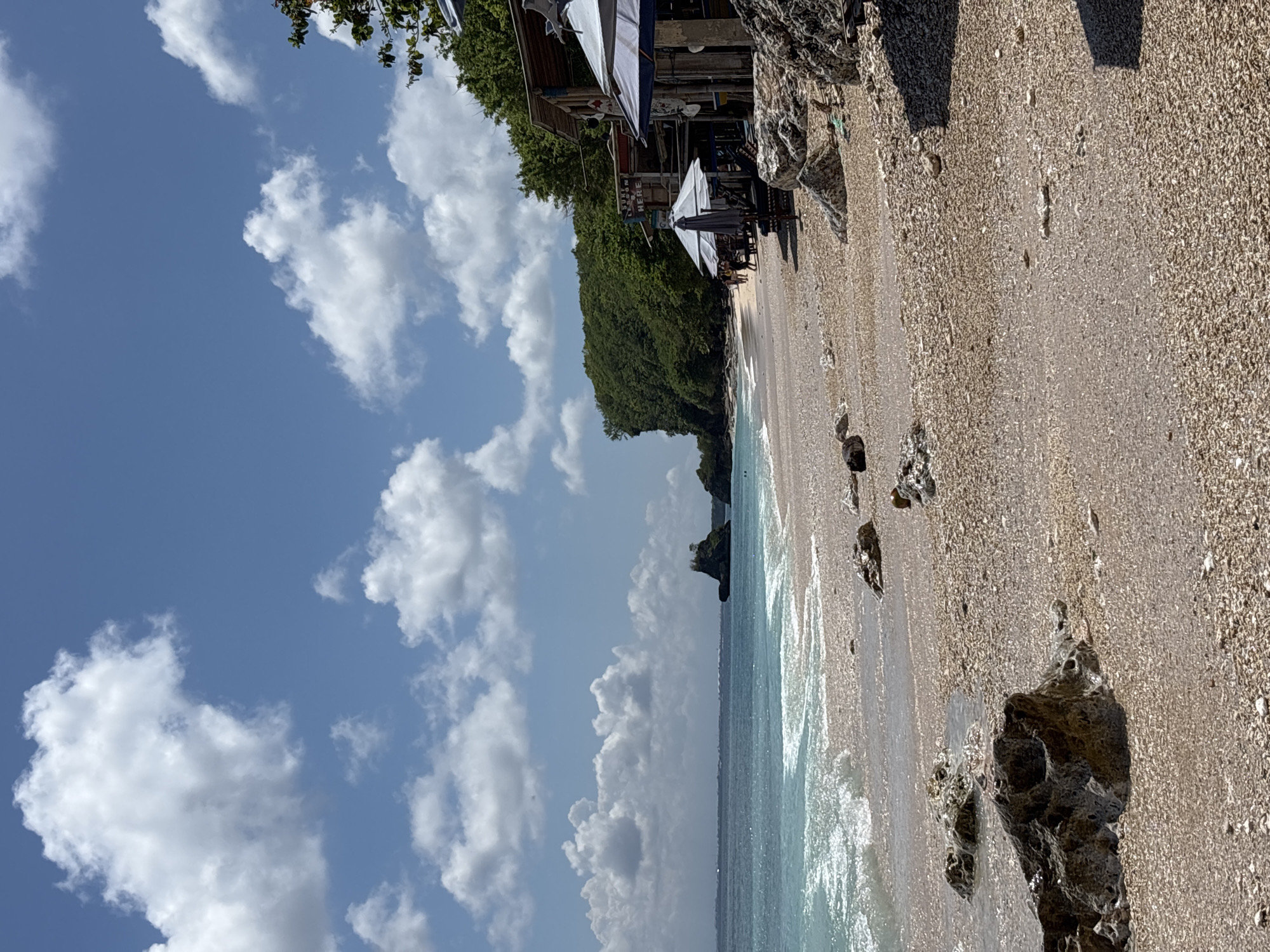 The image depicts a beach scene in Uluwatu, Indonesia. The sky is blue with scattered white clouds. The beach is made of light-colored sand and pebbles, with some scattered dark rocks near the shoreline. The ocean is a light blue-green color, with small waves lapping at the shore. Along the edge of the beach, there is a small restaurant named “SUKON HERE” with parasols and beach chairs. Dense green vegetation covers a cliff or hillside behind the restaurant. There are a few people visible under the umbrellas.