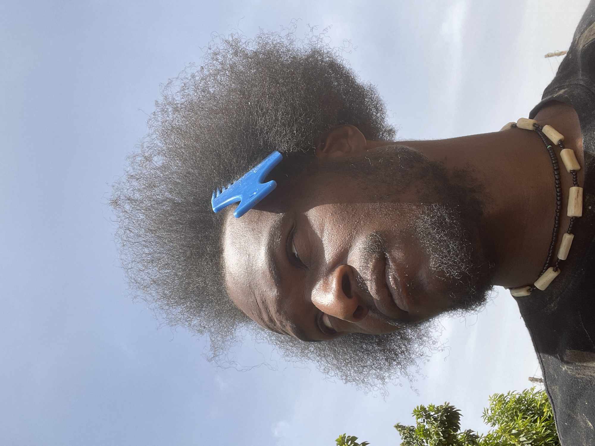 A medium close-up shot depicts a man with a dark complexion and dense, coiled hair. A blue plastic comb is partially embedded in his hair near his forehead. He has a thick, graying beard. He wears a beaded necklace with alternating dark and light-colored beads. The background features a light blue sky. The scene also include some green foliage. While the location is suspected to be in Jos, Nigeria, based on contextual clues, this cannot be definitively determined from the visual content of the image alone.
