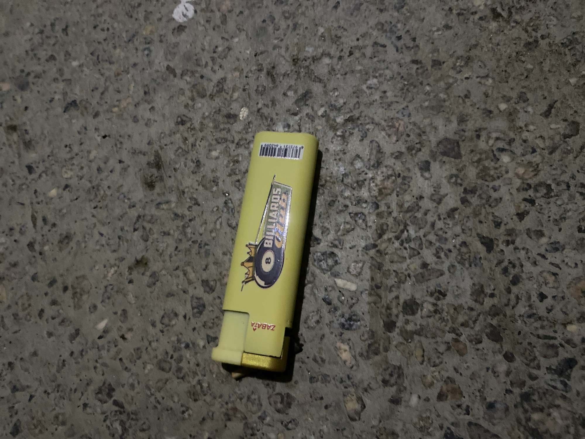 A single yellow (or lime green) lighter is centrally placed on a dark, textured surface, likely concrete or asphalt, exhibiting small embedded stones and general wear. The lighter has a graphic design featuring the word "BILLIARDS" above an 8-ball and a crown-like symbol. On another side, the word "ZABATA" is visible, oriented upside down relative to the main graphic. A barcode with the numbers "979131 042099" is printed near the top of the lighter. The lighting appears dim, suggesting artificial light or low natural light. There are no people or other objects in the frame, and no specific event is taking place. The overall impression is of an object dropped or resting on a ground surface, possibly in Jama A, Nigeria, as provided in the context.