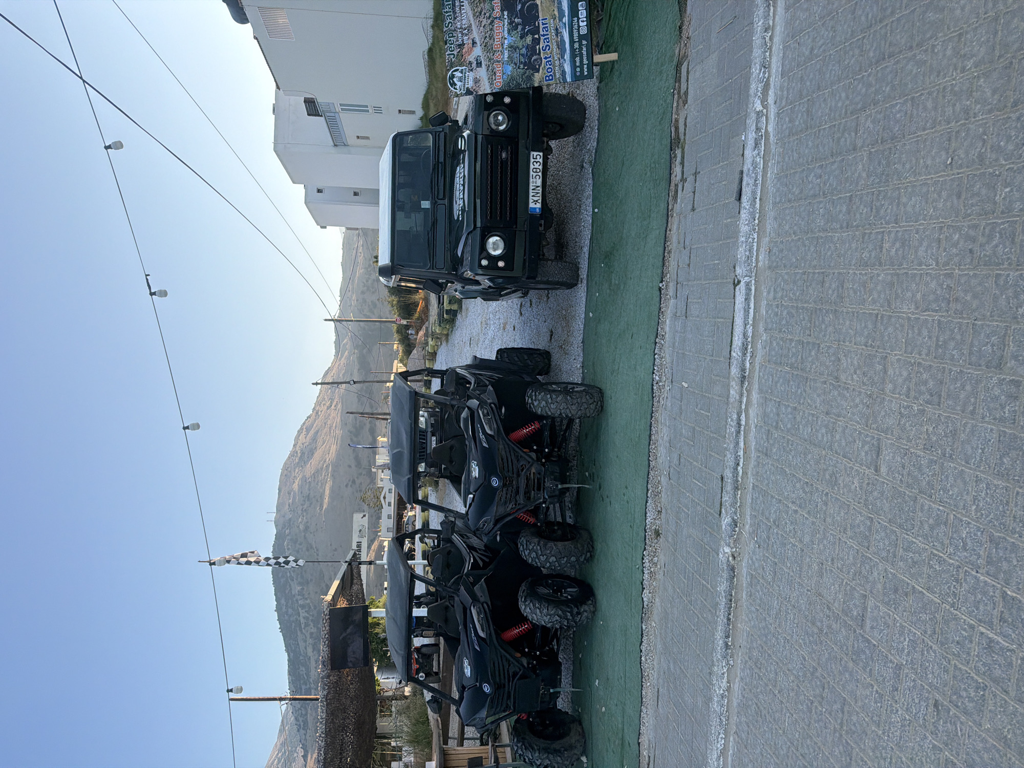 The image depicts a scene with several vehicles parked on a paved area next to a green surface. In the foreground, three black quad bikes are parked in a row. Behind them, a black Land Rover Defender is parked. The vehicles are all facing the same direction.
In the background, there are some buildings and a hillside. A sign advertising "Jeep Safari," "Quad & Buggy Safari," and "Boat Safari" is visible on the building behind the Land Rover. The time of day appears to be late afternoon or early evening, as indicated by the angle of the sunlight and the clear sky. The weather seems to be clear and sunny. The overall setting suggests a location associated with adventure tourism or vehicle rentals, possibly in a coastal or mountainous area given the background. The vehicles are not in motion, and no people are visible, implying the scene is set up for potential customers. The license plate on the Land Rover reads "XNN 5935 GR." A checkered flag is also visible in the distance.