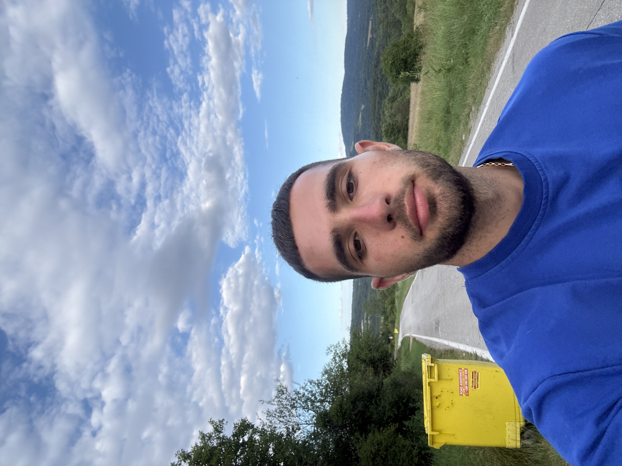 A medium shot captures a young man taking a selfie outdoors. He is positioned in the foreground, facing the camera with a neutral expression. He has short, dark hair and a beard. He is wearing a blue t-shirt. The background depicts a rural landscape with a road winding through rolling hills covered in greenery. A large yellow dumpster is situated to the left of the road. The sky above is partly cloudy, with patches of blue visible. The scene appears to be located on the outskirts of Balinovtsi, Bulgaria, given the presence of a road and natural scenery typical of the Bulgarian countryside.