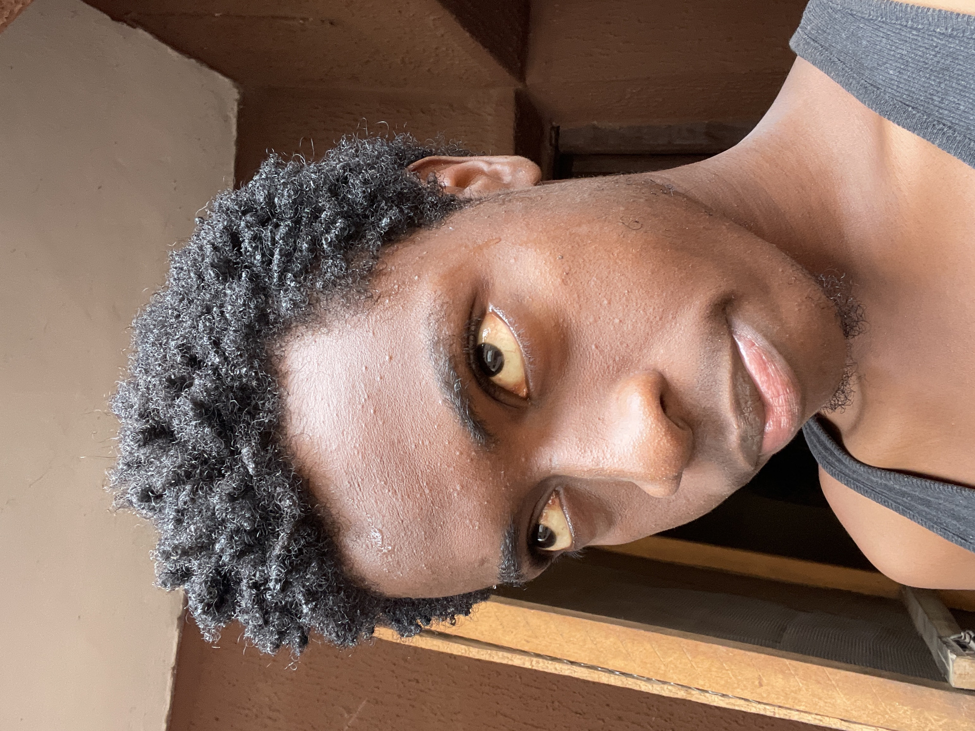 This is a close-up, vertically oriented photograph of a person's face, taken outdoors. The subject, who appears to be a young Black male, is looking directly at the camera with a neutral expression. His skin is dark brown and appears slightly dewy, suggesting sweat or moisture, possibly due to warm weather. He has short, tightly coiled black hair framing his face and a visible ear on the left side of the frame. His eyes are a warm brown, and his lips are slightly parted, revealing a hint of his teeth. The lighting suggests it is daytime.
The background is out of focus, but wooden elements and what appear to be structural beams or a wall are visible, along with a hint of a beige surface. The subject is wearing a dark, possibly black or dark gray, tank top. The overall composition emphasizes the subject's face and features. No text is visible in the image. The provided location context is Nkpor, Nigeria.