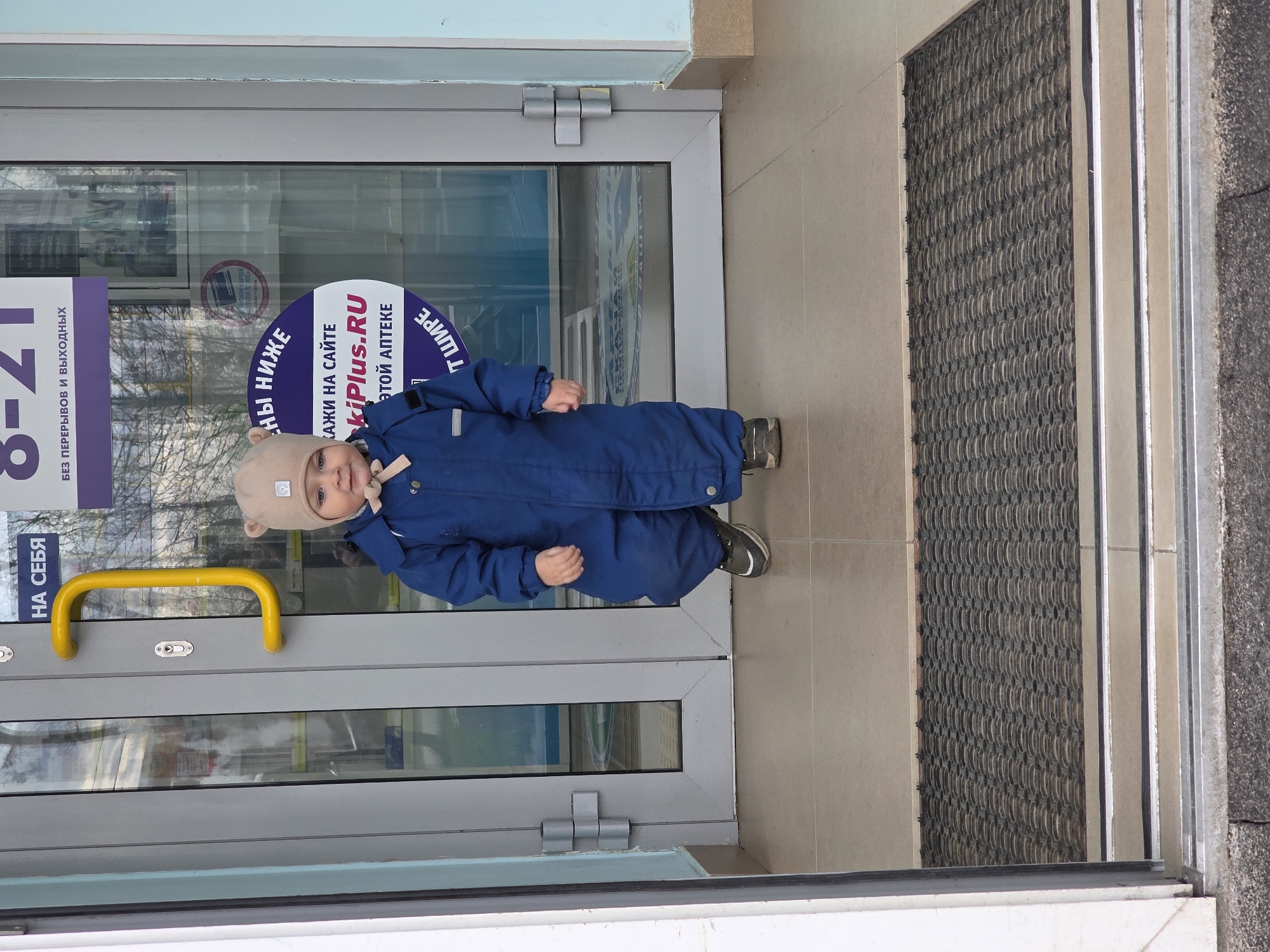 Here is a concise description of the image:
A young child, wearing a blue jumpsuit and a beige hat with bear ears, stands in front of the entrance of a pharmacy. The pharmacy door has a yellow grab bar and signage in Russian indicating the hours of operation and website address. The entrance has a ribbed floor mat. Based on the information on the pharmacy signage, the location is Chaikovsky, Russia.