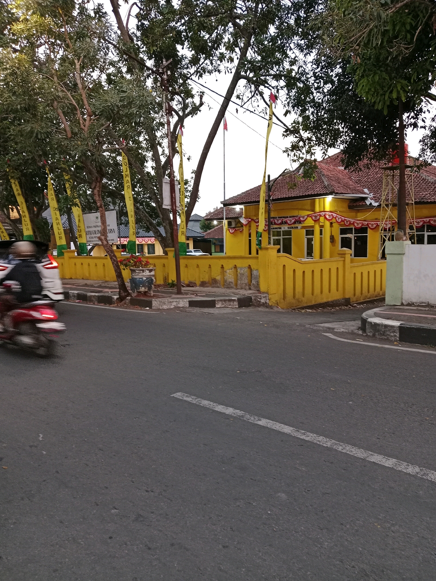The image depicts a street scene in Cianjur, Indonesia, with a yellow building prominently featured in the background. The building has a red tiled roof and is adorned with yellow banners and red and white decorative streamers along its facade. A low yellow concrete wall with vertical indentations runs along the front of the property, separating it from the street. Trees with lush green foliage partially obscure the building and the sky. In the foreground, the asphalt road has a white dashed line marking a lane. A red and white motorcycle with a rider wearing a helmet is in motion on the left side of the road, creating a blur due to movement. There are also cars and a white sign with text in Indonesian visible near the building. The overall lighting suggests it is either early morning or late afternoon.