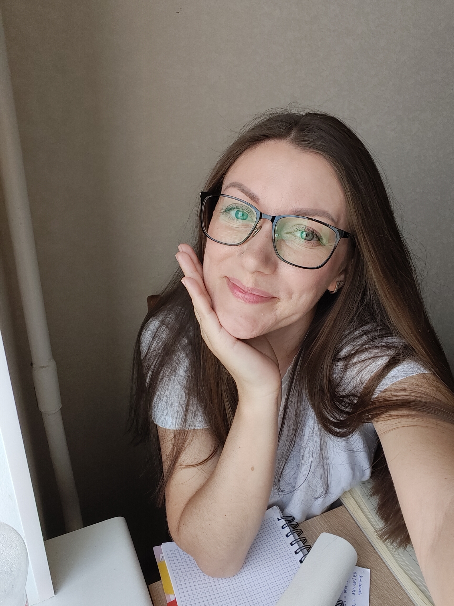 A woman with long brown hair and glasses is resting her chin on her hand. She is wearing a white t-shirt. She is sitting at a desk with a notebook and a roll of paper towel. The background appears to be a wall with a pipe. The image seems to have been taken indoors in Chornomorsk, Ukraine.