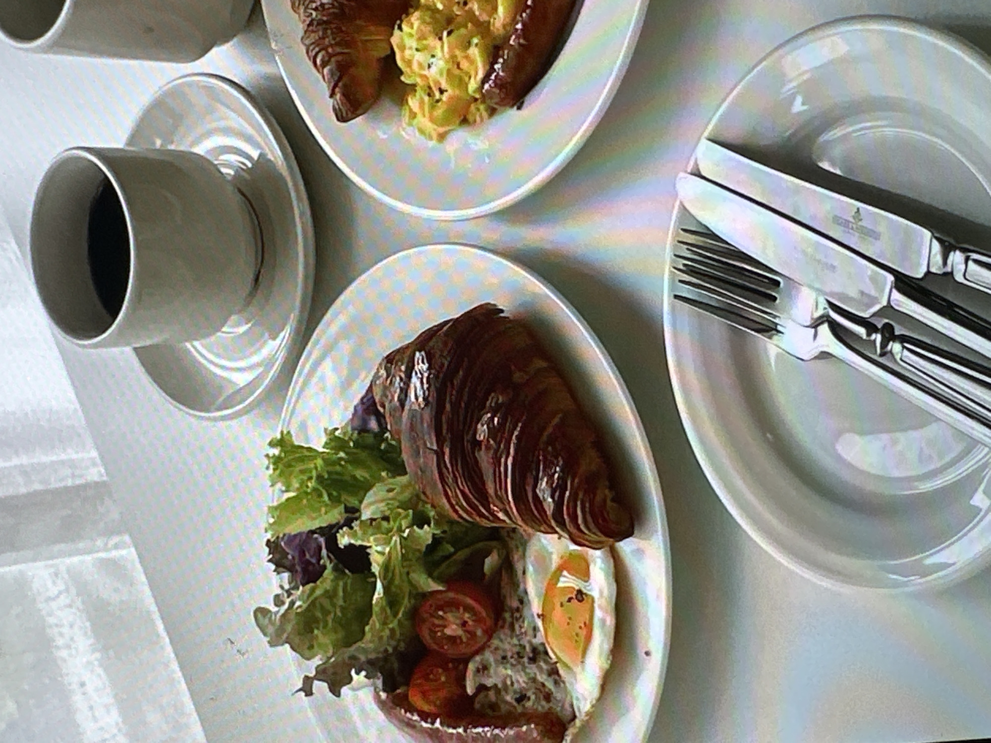 The image presents an overhead shot of a breakfast setting, presumably indoors. There are two white plates. One plate contains a croissant, green lettuce, sliced tomatoes, a sausage, and a fried egg. The second plate contains a croissant, a sausage, and a portion of what appears to be scrambled eggs or a similar dish. A white cup, filled with a dark liquid (likely coffee), sits on a saucer beside the first plate. Adjacent to the second plate is a third white plate, upon which sits a knife and fork. The background is a light-colored surface, possibly a tablecloth. The image does not provide any information about the location in Yerevan, Armenia, or people, subjects, objects, actions, interactions, or the environment beyond what is directly visible.