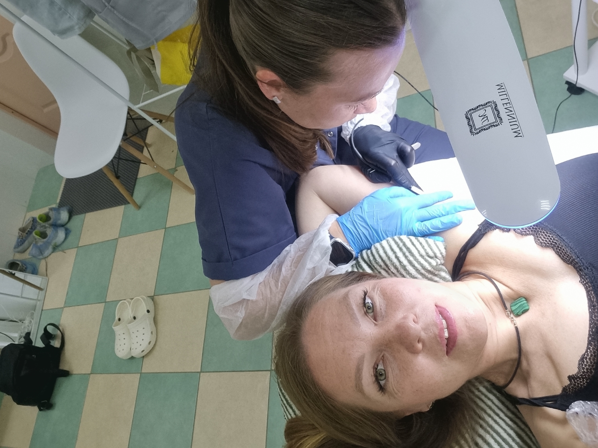 In Chaikovsky, Russia, a close-up shot captures a woman lying down, her face taking up most of the frame. She has light brown hair, green eyes, and is wearing a dark tank top with lace trim and a necklace with a green stone pendant. Another person, partially visible from the shoulders down, is leaning over her, wearing a dark blue top and disposable gloves, one of which is light blue and the other black. This person appears to be working on the woman's arm, near a white LED lamp with a logo that reads "MILLENNIUM" in reverse. The environment is a room with a tiled floor in a checkerboard pattern of light green and cream squares. In the background, a white chair with wooden legs and a black backpack are visible.