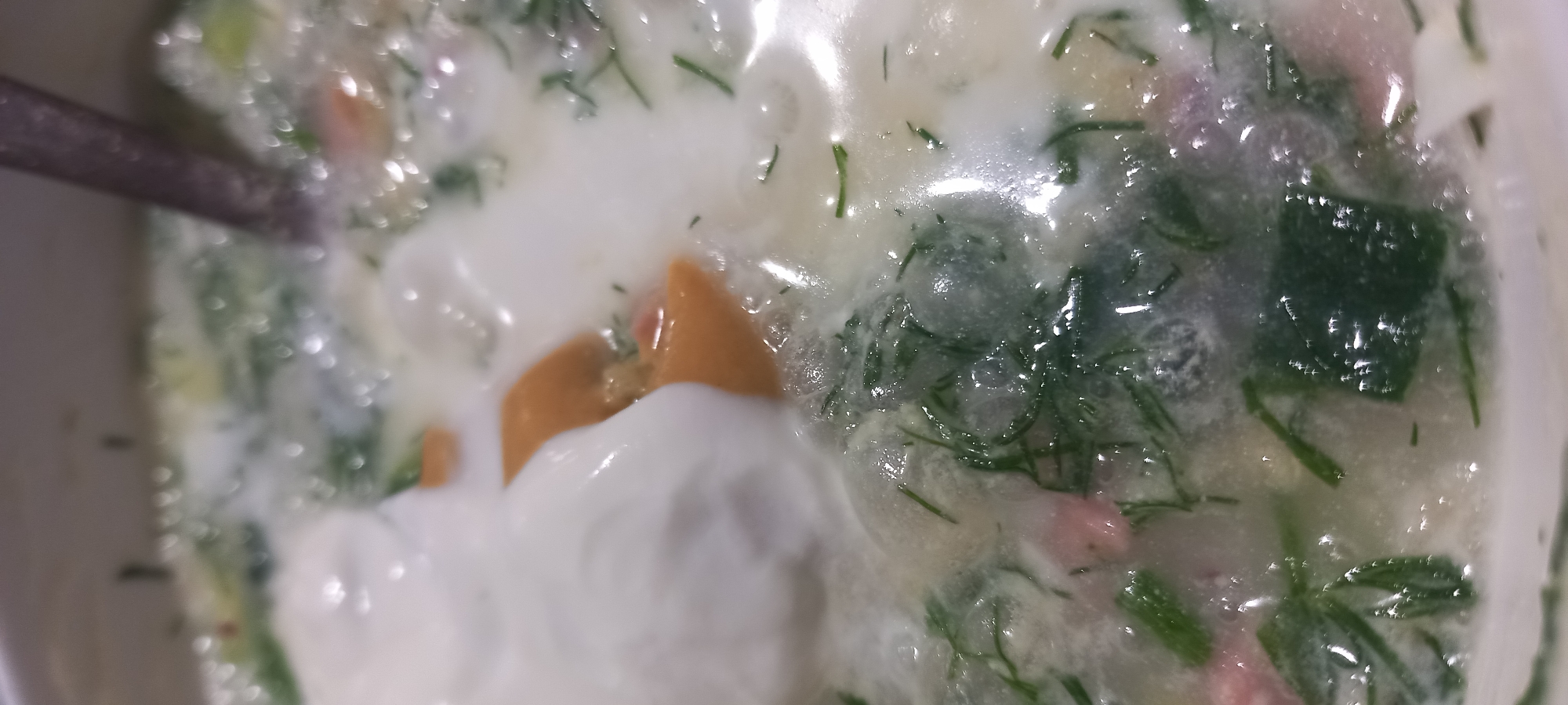 The image shows a close-up view of a bowl of soup. The soup appears to be a cold soup, possibly Okroshka, with ingredients such as chopped dill, what looks like boiled eggs, and possibly meat or vegetables. A large dollop of sour cream or yogurt is visible on top of the soup. The surface of the soup is bubbly and slightly frothy, with a spoon visible on the right side of the frame. The lighting is bright, creating highlights on the liquid surface. There is no identifiable information in the image that would suggest its location in the city of Kryvyi Rih, Ukraine.
