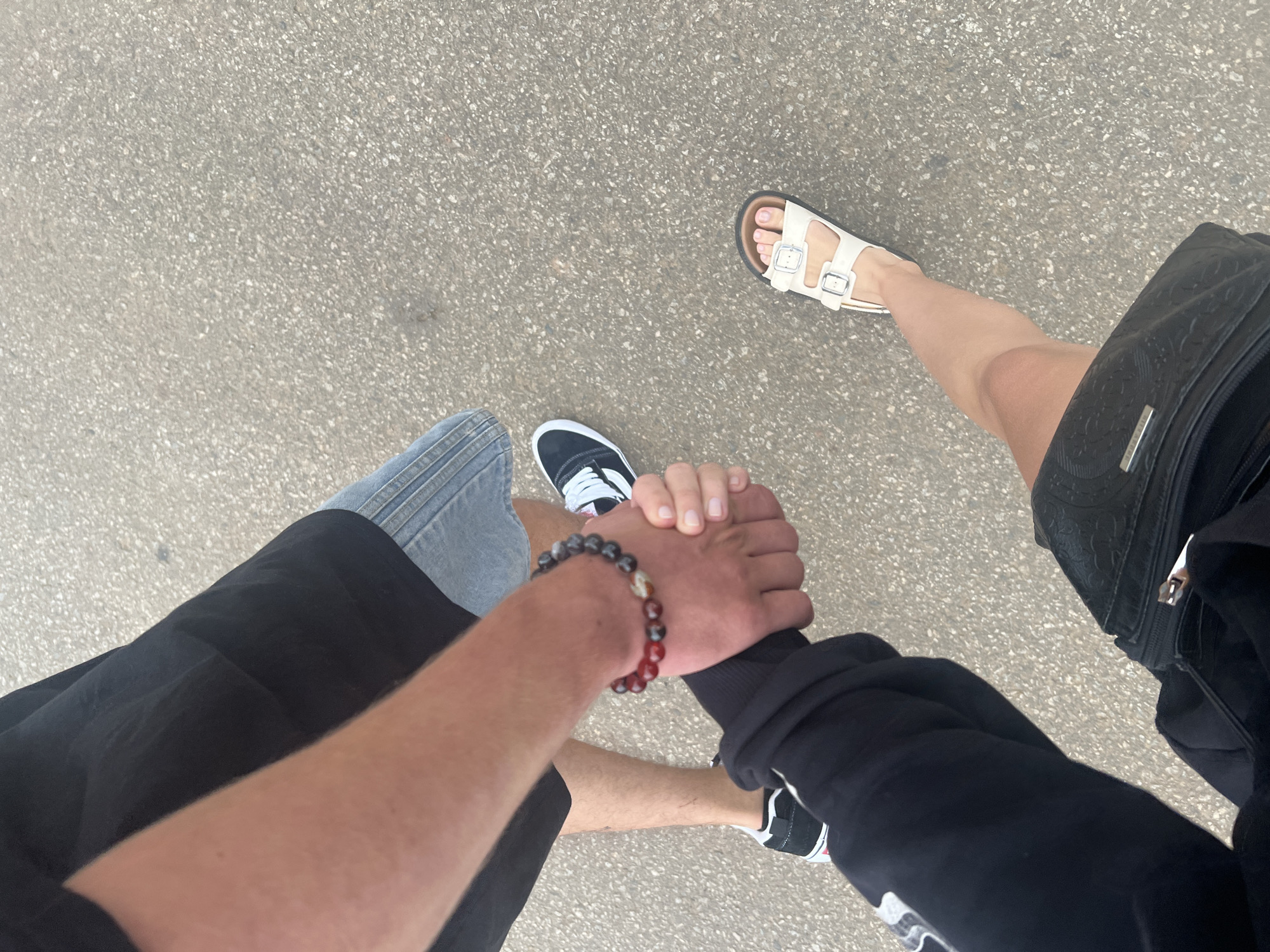 An overhead photograph captures two people holding hands while standing or walking on a light grey asphalt surface in Bo Y, Vietnam. The perspective looks down, primarily showing their lower bodies, feet, and clasped hands.
On the left side of the frame, one individual is seen wearing light blue denim shorts and black sneakers with white laces and soles. Their arm is covered by a black long-sleeved garment, and a beaded bracelet with a mix of red and dark, possibly agate, beads adorns their wrist.
The person on the right wears black shorts or a skirt and white, two-strap sandals. A black bag with a subtle patterned texture and a small metallic, rectangular embellishment is visible against their leg. Both individuals' legs appear bare.
Their hands are firmly clasped together in the center of the image, suggesting a connection, companionship, or affection between them. The ground is a typical speckled grey pavement, indicating an outdoor setting during the daytime. No specific time of day or weather conditions are explicitly discernible, but the natural light suggests clear conditions. No text is clearly readable on any items.