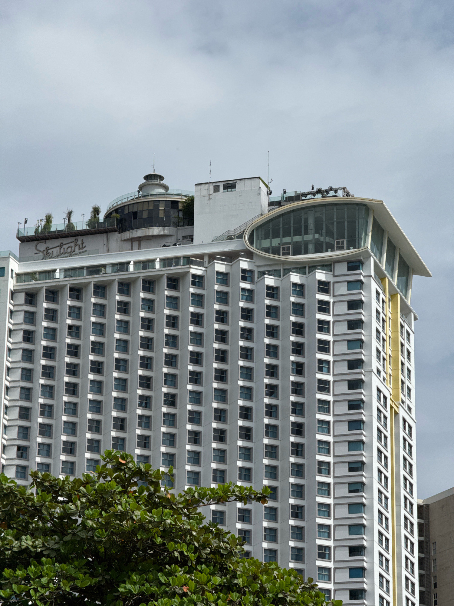 This is a medium-angle, eye-level shot of the Skylight Nha Trang, a multi-story modern building located in Nha Trang, Vietnam. The building features a white facade with numerous rectangular windows arranged in a grid pattern. The top of the building has a distinctive curved glass structure and a rounded rooftop area with a central circular element, possibly an observation deck or restaurant. There is a prominent "Skylight" sign on the upper facade. Lush green foliage from a large tree partially obstructs the lower portion of the building. The sky is overcast and cloudy.