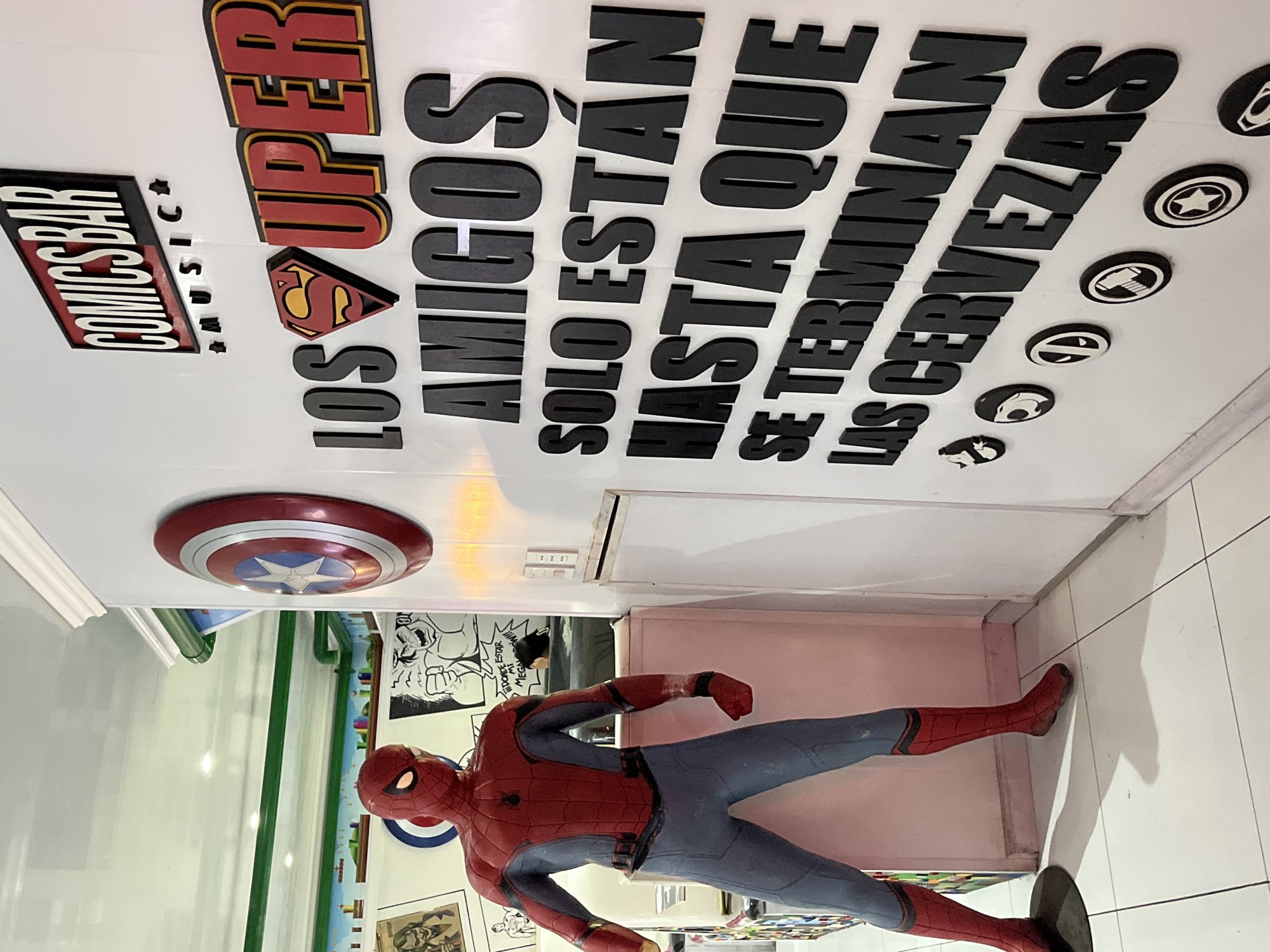 The image shows the interior of a bar decorated with comic book-themed elements. A Spider-Man statue lies on the floor. Above it, on the ceiling or a high wall, are signs reading "COMICS BAR MUSIC" and "LOS SUPER AMIGOS SOLO ESTÁN HASTA QUE SE TERMINAN LAS CERVEZAS". There is also a Captain America shield and other comic book symbols. The location is Unknown, Unknown.
