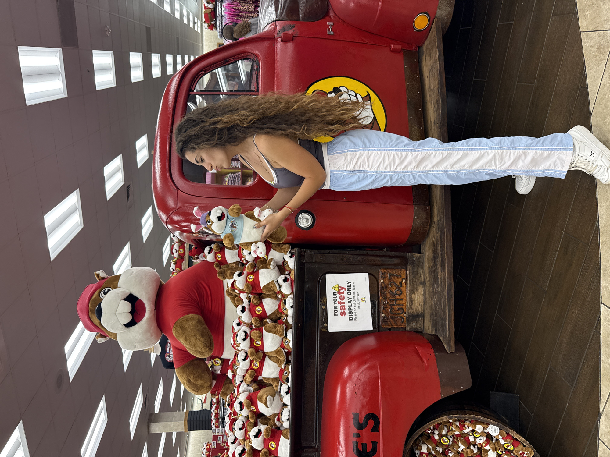 An interior photograph depicts a young adult female standing next to a red vintage-style pickup truck merchandise display within a Buc-ee's travel center in Fort Valley, Georgia, United States. The female, possessing long, dark, curly hair, is attired in a dark grey tank top, light blue track pants with vertical white stripes, and white athletic shoes. She holds a light-colored Buc-ee's beaver mascot plush toy, wearing a light blue shirt and bunny ears, in both hands, looking towards it with pursed lips. The bed of the red truck is filled with numerous smaller Buc-ee's beaver plush toys, each wearing a red shirt. A large plush Buc-ee's beaver mascot figure, dressed in a red shirt and cap, is positioned behind the truck bed. A white sign affixed to the truck bed reads, "FOR YOUR SAFETY DISPLAY ONLY Please do not climb on, lean on or sit in truck." Below the sign, a brown Texas license plate bearing "JGH627" is visible. A wooden barrel overflowing with small Buc-ee's plush toys is located near the truck's front wheel. The store environment features dark wood-patterned floor tiles and a ceiling equipped with rectangular fluorescent lighting fixtures. Additional retail displays are discernible in the distant background.