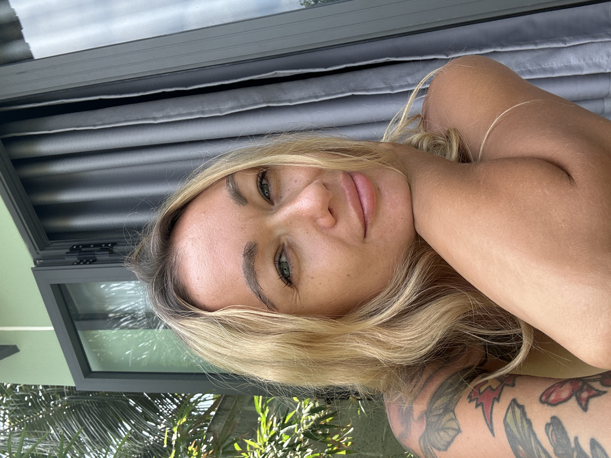 In Mũi Né, Vietnam, a fair-skinned woman with blonde hair and green eyes rests her chin on her arms. She is wearing a light-colored top, and her left arm features a colorful tattoo. The background shows a light green wall with a large glass window and gray curtains. Lush green foliage is visible to the left of the frame, suggesting an outdoor setting. The lighting indicates it is daytime.