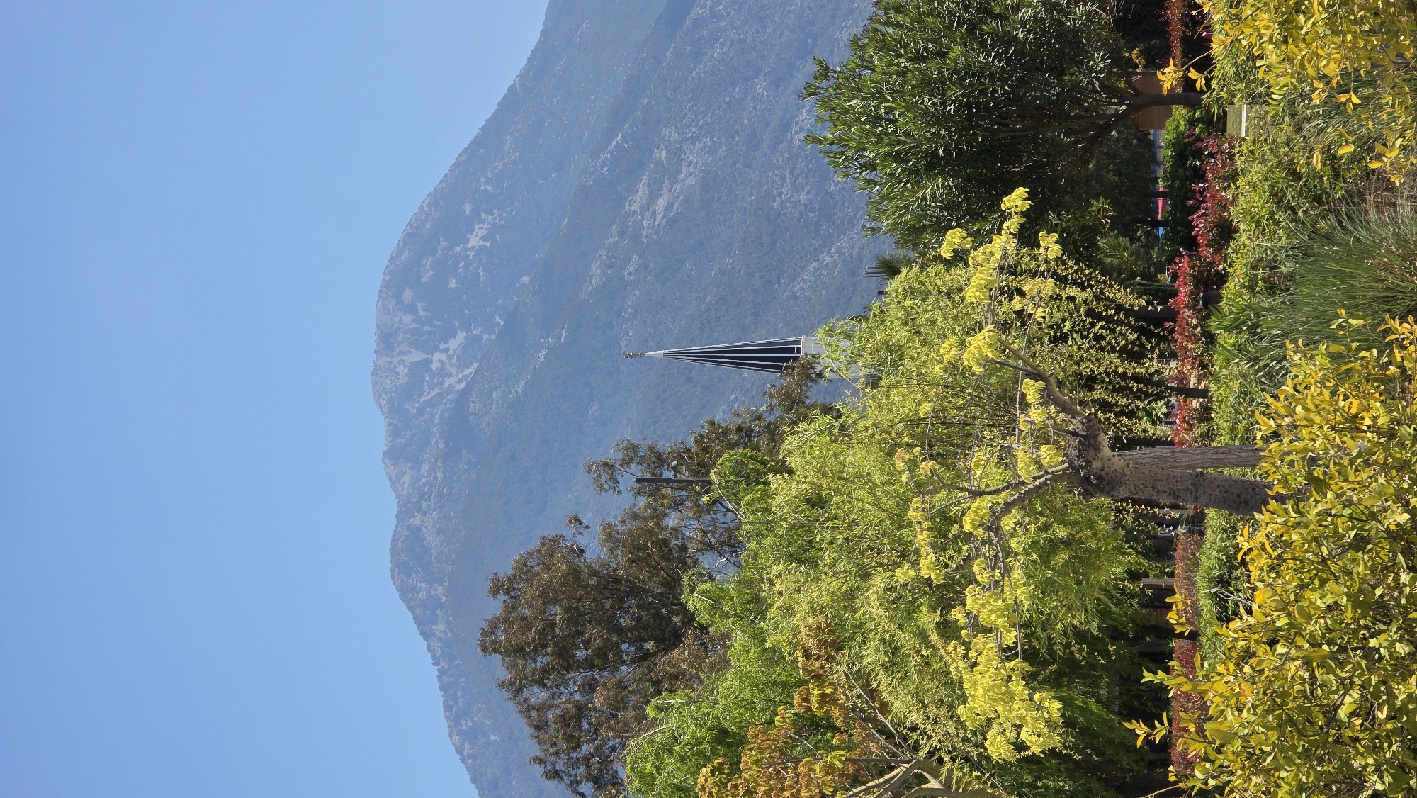 The image depicts a view of a mountain and vegetation with a spire partially visible through the trees. The mountain is light blue and has a rugged texture. The trees in the foreground are a mix of green and yellow hues. The spire is black and silver and appears to belong to a church or similar structure. This is likely a view of the mountains near Fethiye, Turkey, and part of the Fethiye Church, based on its spire.
