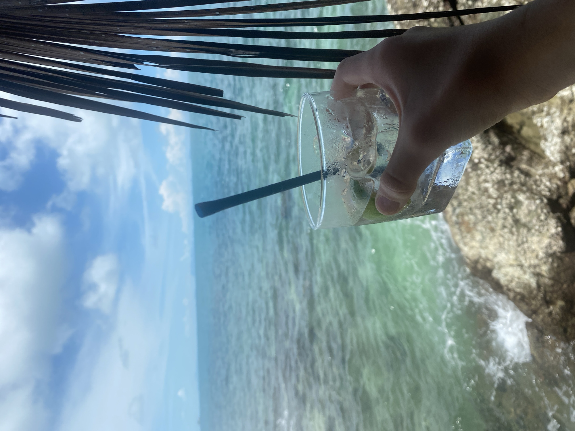 A hand holds a clear glass filled with ice and a light-colored beverage, a dark straw inserted. The glass is positioned in the foreground, with the backdrop showcasing a bright, partly cloudy sky and a turquoise ocean. The ocean waves gently crash against a rocky shore in the lower portion of the image. On the right side, a portion of a dried palm frond is visible, obscuring a small section of the view. The overall impression is a serene, tropical setting, likely on Koh Phangan, Thailand.