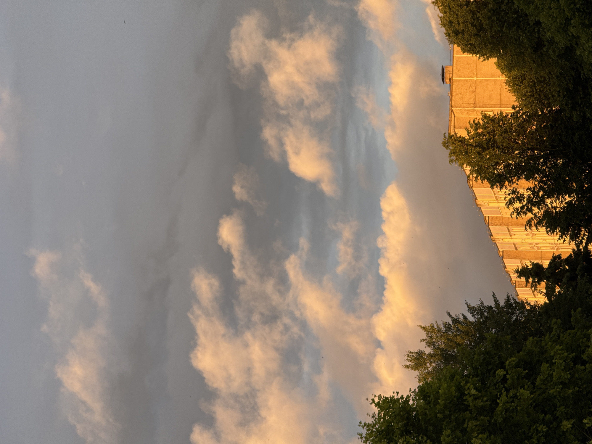 A low-angle, outdoor, full shot captures the late afternoon sky above a multi-story, light-orange apartment building. The sky is filled with a mix of heavy, dark grey clouds and lighter, wispy clouds illuminated by the golden light of the setting sun. The building, which occupies the lower right portion of the frame, is partially obscured by dark green trees. A chimney stack is visible on the roof of the building. The image appears to have been taken in a residential area of Minsk, Belarus, with no individuals or overt actions depicted.