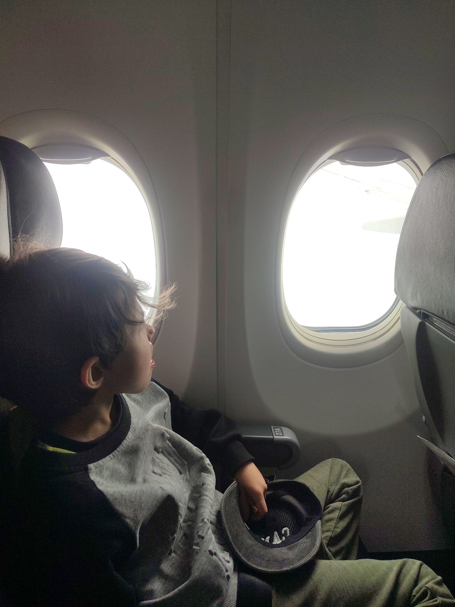 A young boy sits by the window of an airplane, looking out. He has short, dark hair and is wearing a two-toned sweatshirt. In his lap, he holds a cap. The airplane window is bright, with a view of the wing. The seat is upholstered in dark grey. The seat number "21A" is visible on the armrest. It is not possible to determine a location or city based on the image.