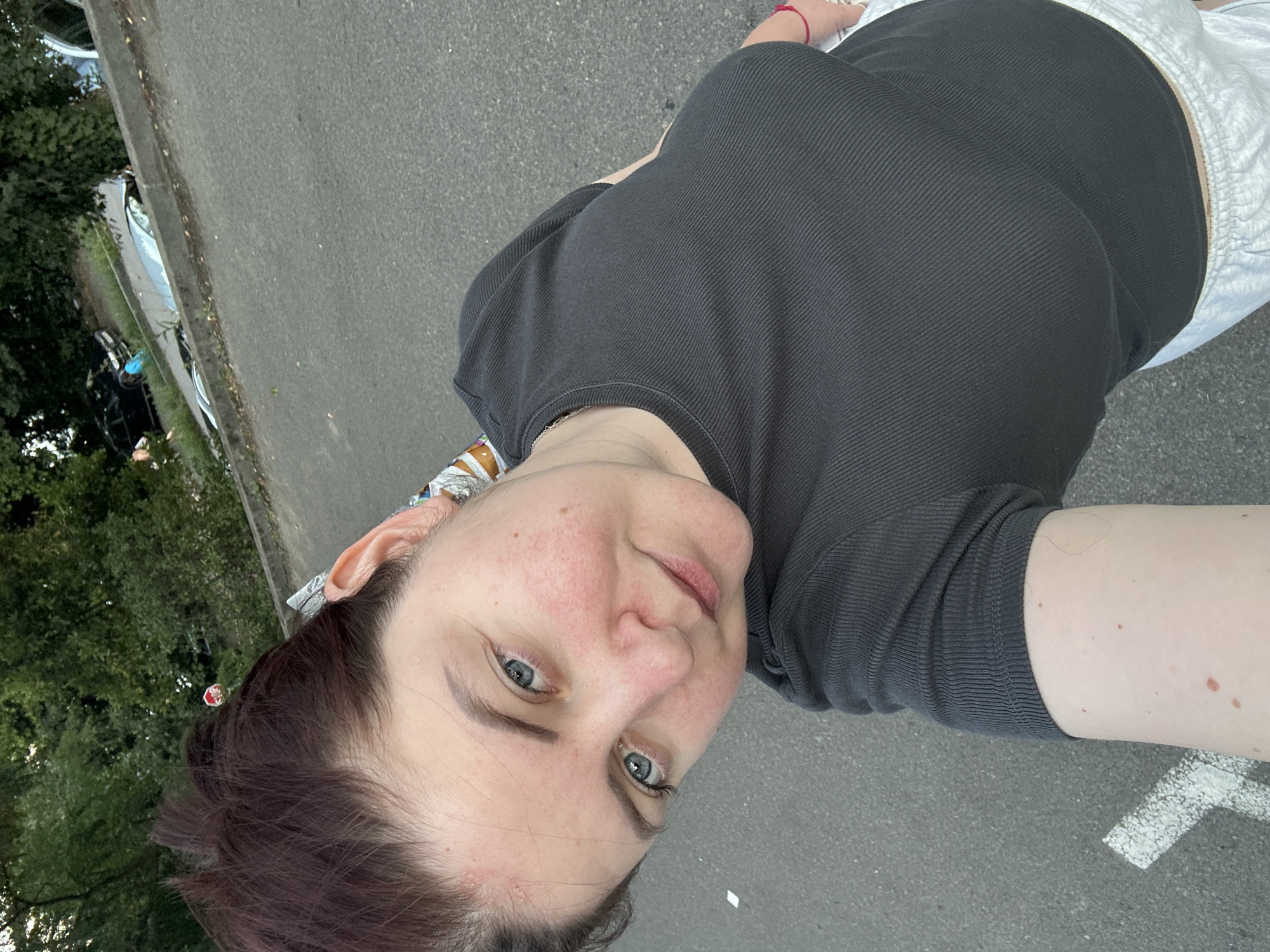 The image is a selfie taken outdoors, likely in a parking lot or alongside a road. The subject is a woman with auburn hair pulled back, wearing a dark grey ribbed t-shirt and light-colored shorts. She has blue eyes and fair skin with some redness on her cheeks. In the background, trees and parked cars are visible. The ground is asphalt with white markings. The specific location in Lovran, Croatia, is not discernible from the visual information in the image.
