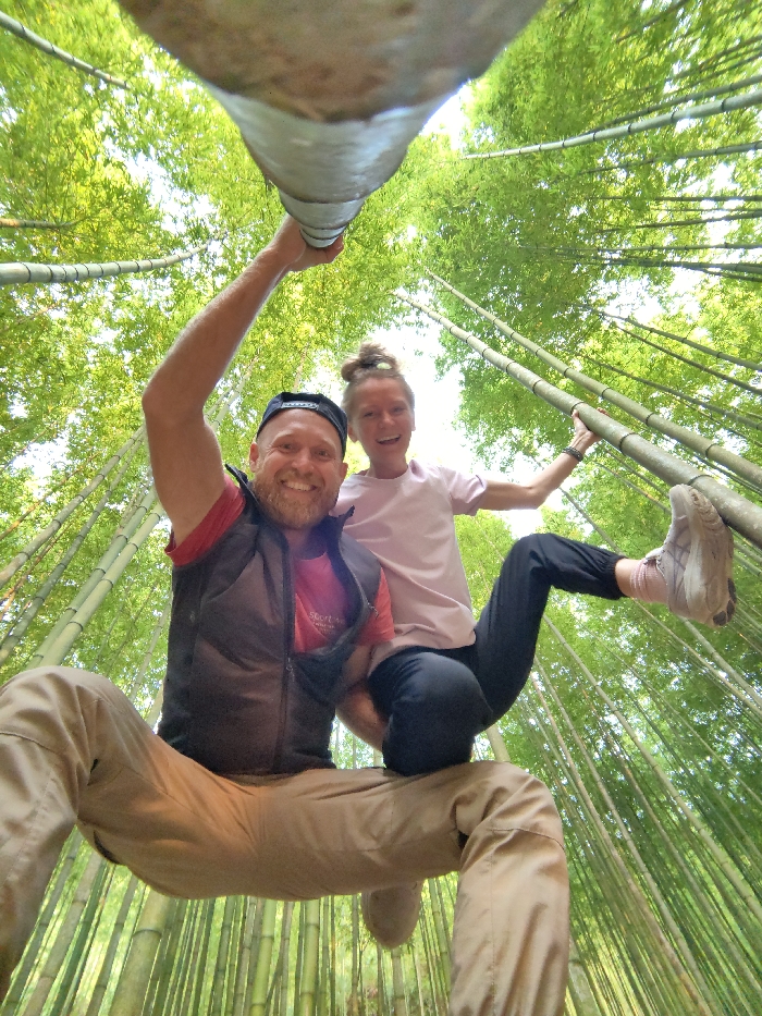 A low-angle, wide-lens photograph depicts two individuals, one male and one female, within a dense bamboo forest in Mù Cang Chải, Vietnam. Both subjects are smiling and looking directly at the camera. The male, positioned on the left, wears a backward black baseball cap, a red shirt, a dark sleeveless vest, and khaki pants. He extends his left arm upwards, grasping a thick, light-colored horizontal bamboo pole above his head, appearing suspended with his legs bent and splayed. The female, on the right, has her light hair tied in a bun and wears a light pink t-shirt, dark leggings, and light athletic shoes with pink socks. She holds a vertical green bamboo stalk with her right hand and places her left foot against another bamboo stalk, suggesting a climbing or balancing pose. The background consists entirely of numerous tall, slender green bamboo culms and their vibrant green foliage, extending upwards towards a bright, unseen sky. The scene is illuminated by natural light, indicating an outdoor, daytime setting.