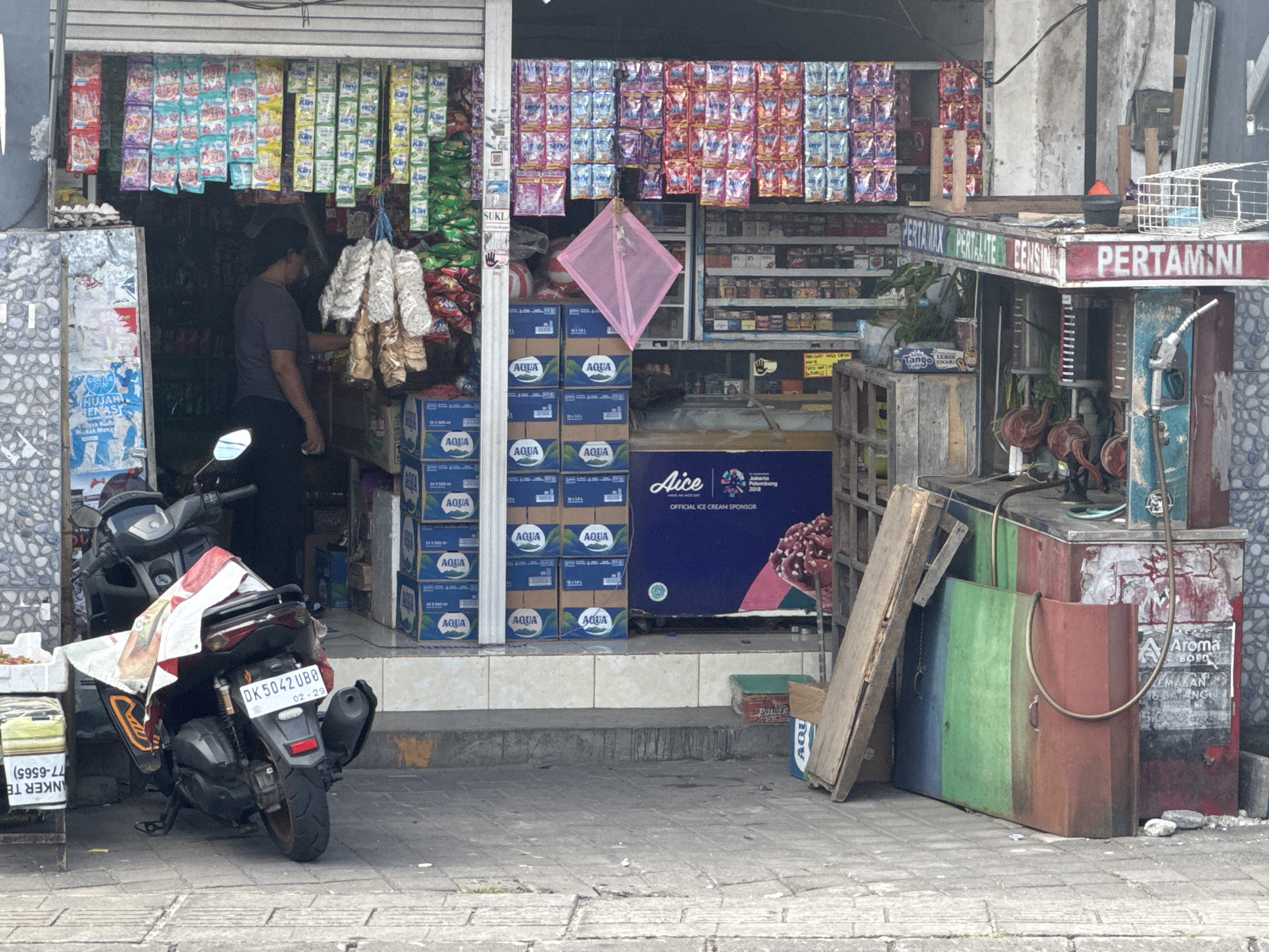 The image captures an outdoor scene in Kuta Selatan, Indonesia, depicting a small shop with goods displayed both inside and outside. A person, wearing a dark shirt and cap, is visible inside the shop, reaching towards hanging items. In front of the shop, a dark-colored motorcycle with a license plate reading "DK 5042 UBB" is parked. Several boxes of "AQUA" bottled water are stacked near the entrance. A prominent sign above a fuel pump reads "PERTAMINA" and "PERTALITE EENSIN". To the right of the fuel pump, there are several large metal containers, one red, one green, and one with a brown hue, with wooden planks leaning against them. The shop's facade is adorned with numerous hanging packets of snacks and other products. A pink kite is suspended from the awning. The ground is paved with grey stone tiles.