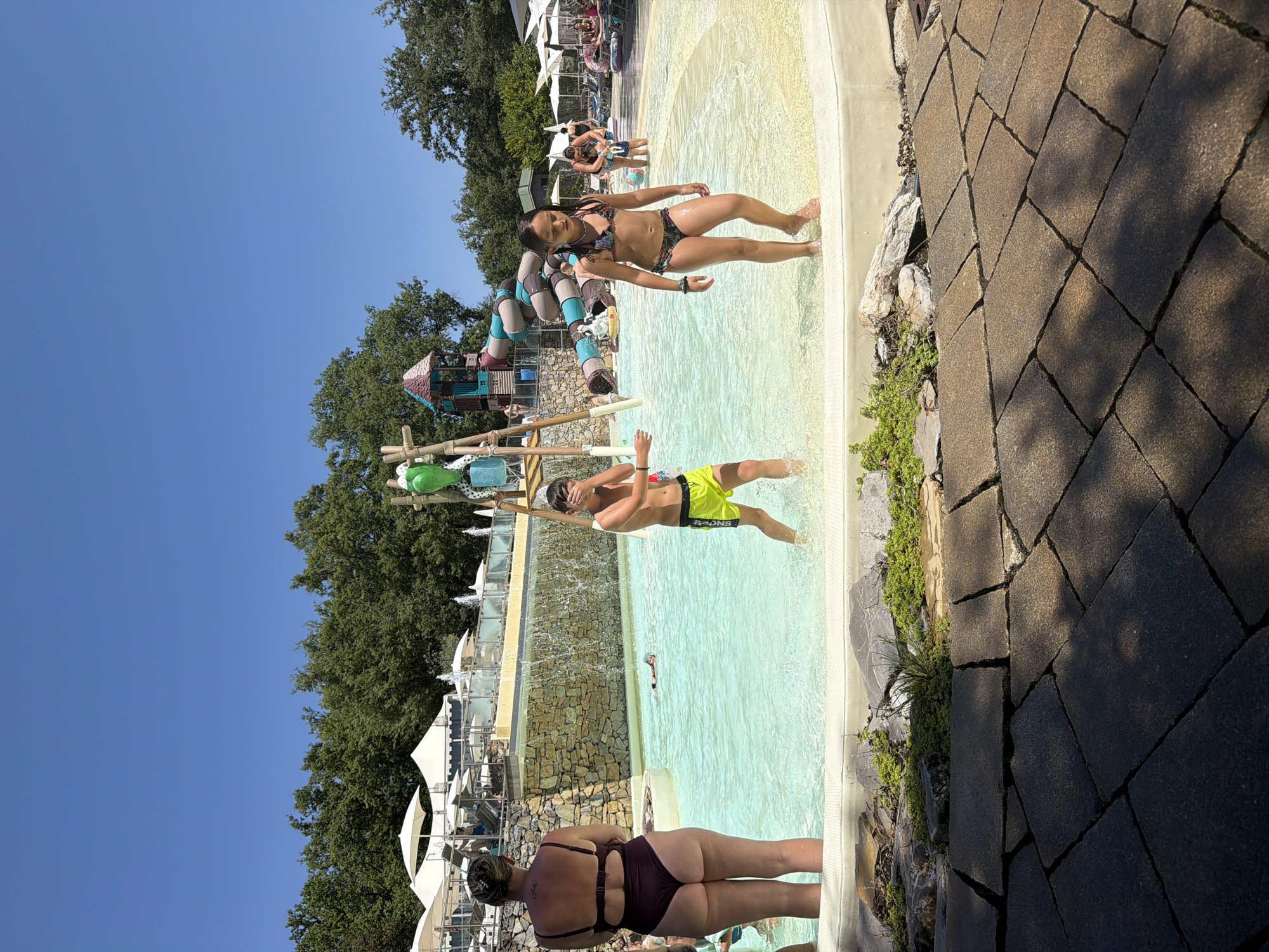 This media file depicts an outdoor water park on a sunny day. The scene features a shallow pool area with a woman in a dark-colored one-piece swimsuit standing at the edge of the pool. In the pool, a young boy in neon yellow swim trunks is playing, with water splashing around him. Further in the background, a young woman in a colorful bikini is posing near the pool edge.
The water park has various attractions, including a colorful water slide structure with tubes and climbing elements, visible in the mid-ground. There are also white umbrellas providing shade for lounge areas in the background, suggesting a leisure and recreational setting. The pool area is bordered by light-colored paving stones and natural elements like rocks and greenery. The sky is a clear, bright blue, indicating good weather. The overall atmosphere appears to be one of enjoyment and summer fun.
There is no visible text in the media file. The location context provided indicates Cavriglia, Italy. The time of day appears to be daytime, likely mid-afternoon given the lighting and shadows.