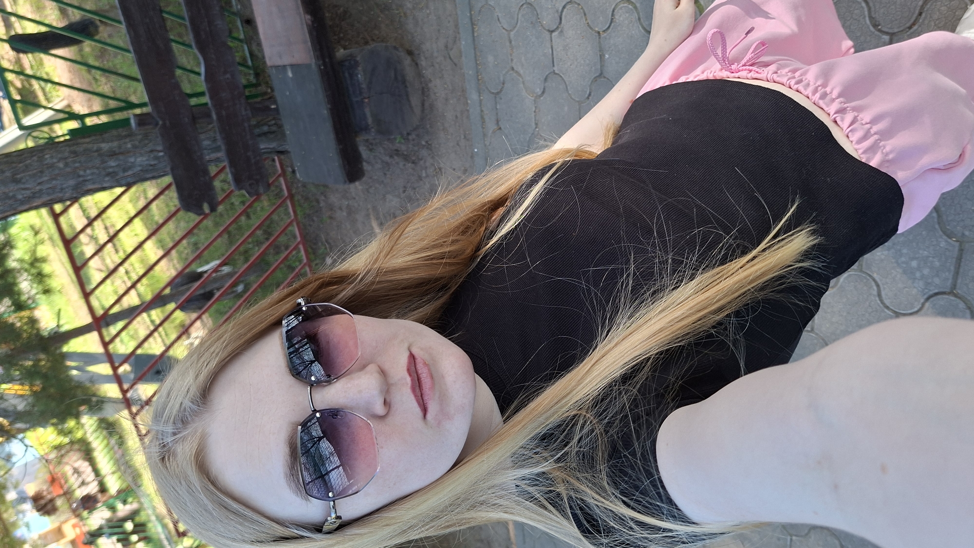 A young woman with long blonde hair and sunglasses is taking a selfie outdoors. She is wearing a black top and pink pants with a drawstring. The background includes a park-like setting with trees, a fence, and a paved area. Based on the information provided, the location is Chaikovsky, Russia.