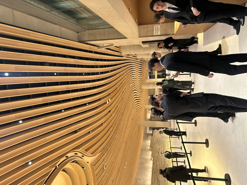 The image depicts the interior lobby of the Philharmonie de Paris, located in the 19th arrondissement of Paris, France. The scene is dominated by a distinctive, undulating ceiling composed of numerous parallel wooden slats of light wood, curving organically across the expansive space and featuring integrated spotlights. The floor is light-colored and polished.
On the left side of the frame, a textured wall reveals exposed ancient stone structures, possibly archaeological ruins, behind a series of black stanchions with retractable belts. Several individuals are present in this area; one male in a dark suit stands looking towards the ruins, while another male in a dark jacket walks away from the viewer.
In the mid-ground and foreground, a group of approximately 10-15 individuals, predominantly young to middle-aged males, are gathered. Many are dressed in dark suits, suggesting a formal event. Two males in dark suits are engaged in conversation at the center-right, one gesturing with his right hand. To their right, three additional males in dark suits are visible, one on the far right facing the viewer directly. Other individuals in suits and casual attire are scattered in the background, some appearing to be staff. A light-colored pillar on the right features a rectangular sign with text. Interactions primarily involve standing and conversing within small groups.