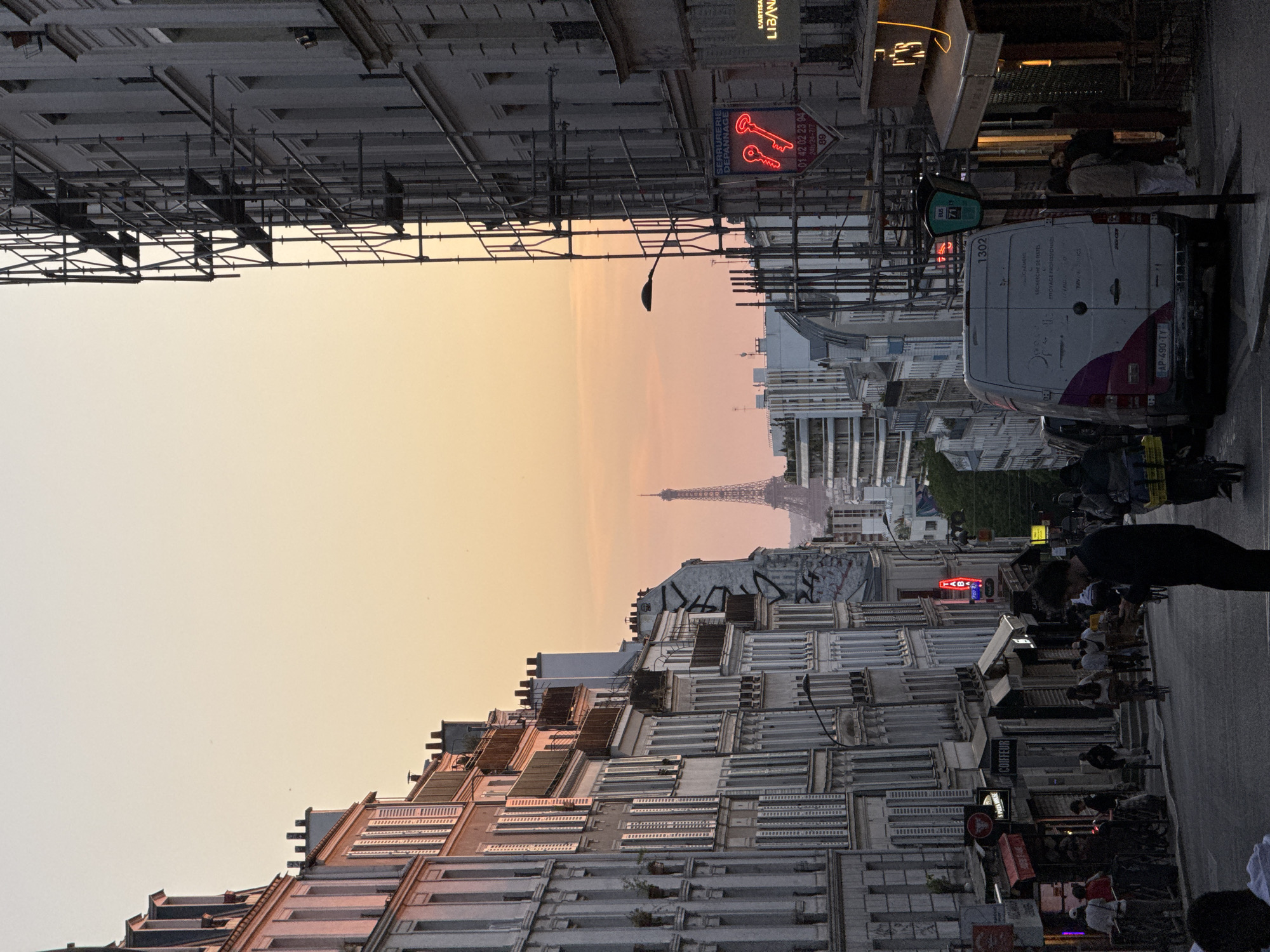 The image presents a vibrant street scene in Paris, France, captured during the golden hour as dusk settles. The perspective looks down a bustling street lined with multi-story buildings, culminating in a clear view of the distant Eiffel Tower silhouetted against the colorful sky. The sky transitions from a soft orange and pink at the horizon to a paler yellow and blue higher up, indicating a clear evening.
On the left, a tall building is partially covered with extensive scaffolding, suggesting ongoing construction or renovation. The street below is active with several people; some are walking, while others appear to be seated at outdoor tables of cafes or restaurants, enjoying the evening. A person in a dark shirt is prominent on the right side of the frame, facing away from the viewer.
Various objects of urban life are visible, including streetlights, a traffic light, and a green bus stop sign marked "BUS 74". A white Mercedes-Benz Sprinter van is parked on the right, displaying "1302" on its rear door and the license plate "AP-490-TY". The van also has text such as "RECHERCHE DE FUITES" (leak detection) and "PROYAGE PROFESSIONNEL" (professional cleaning/spraying).
Storefronts along the street are illuminated with artificial lights and neon signs. A distinctive red neon sign, shaped like two keys, advertises "SERRURERIE DÉPANNAGE" (locksmith service), along with a phone number "01 42 02 23 94" and "24h/24 - 7/7" availability. Other visible text on businesses includes "L'ARTISAN LIBAN," "COIFFEUR" (hairdresser), and "TABAC" (tobacco shop). The overall atmosphere is lively and picturesque, capturing the essence of Parisian street life at sunset.