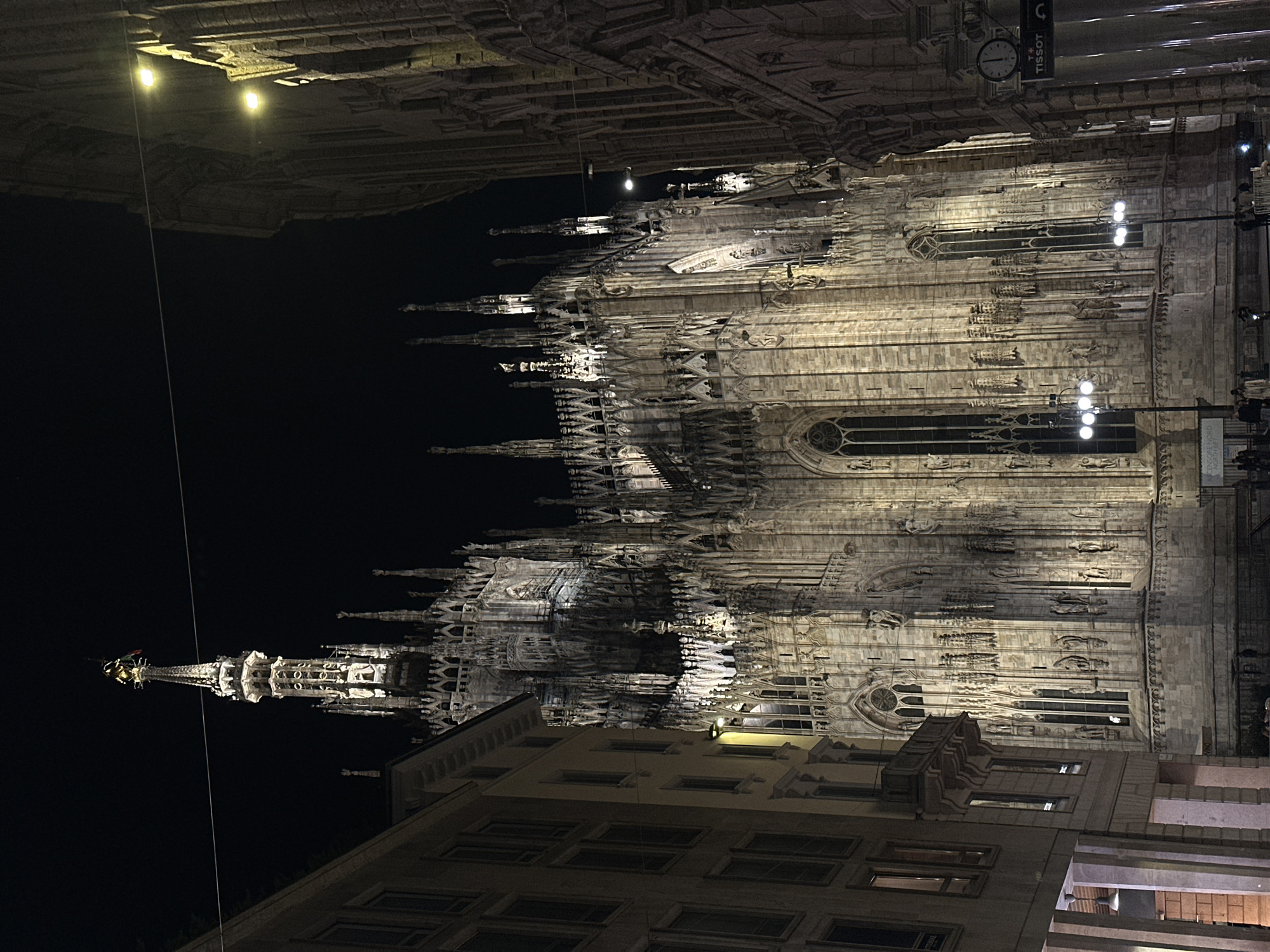 The image depicts the Milan Cathedral (Duomo di Milano) at night, viewed from a low angle. The cathedral's facade is illuminated, showcasing its intricate Gothic architecture, including spires, statues, and windows. A building is visible in the foreground. A Tissot clock can be seen in the upper right. The sky is black.