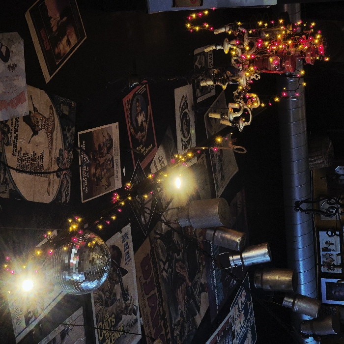 The scene depicts the ceiling and upper wall area of an interior space, specifically located at Le Graffalgar hotel in Strasbourg, France. The dark ceiling is extensively covered with numerous vintage movie posters, many appearing aged or distressed, featuring films such as "Jaws" and "Casino Royale." A silver-faceted disco ball hangs centrally, illuminated by a bright, direct light source positioned above it. Strands of small, multi-colored LED string lights, primarily red and yellow/orange, are strung across the ceiling, draping over various objects. Several silver-toned metal canisters or small milk churns of varying sizes are suspended from the ceiling by wires. To the right, a multi-armed chandelier, possibly made of a light-colored material, is heavily intertwined with the same red and yellow/orange string lights, obscuring some of its details and creating a vibrant glow. Another bare, bright light bulb is visible among the hanging decorations. Below these ceiling elements, a large, horizontal, silver-colored ventilation duct or pipe is visible. Further down on the dark wall, several framed items or pictures are dimly discernible. No individuals are present in the frame, and no specific actions or interactions are depicted. The environment is characterized by eclectic, themed decor and ambient lighting from the various fixtures.