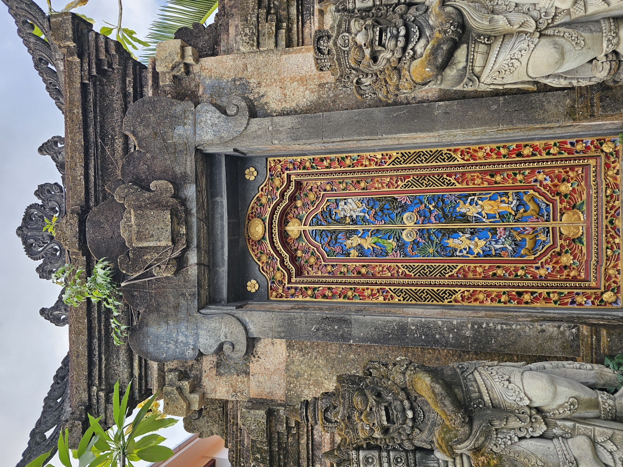 The image shows a traditional Balinese temple gate in Denpasar, Indonesia. The gate features two imposing stone statues of Dwarapalas, guardian deities, flanking a ornately carved wooden door. The door is painted in vibrant colors, with intricate designs of figures and floral motifs. The temple structure itself is made of stone, with weathered textures and some vegetation growing on its surface. The sky is overcast, suggesting a cloudy day. The overall scene evokes a sense of ancient culture and spirituality.