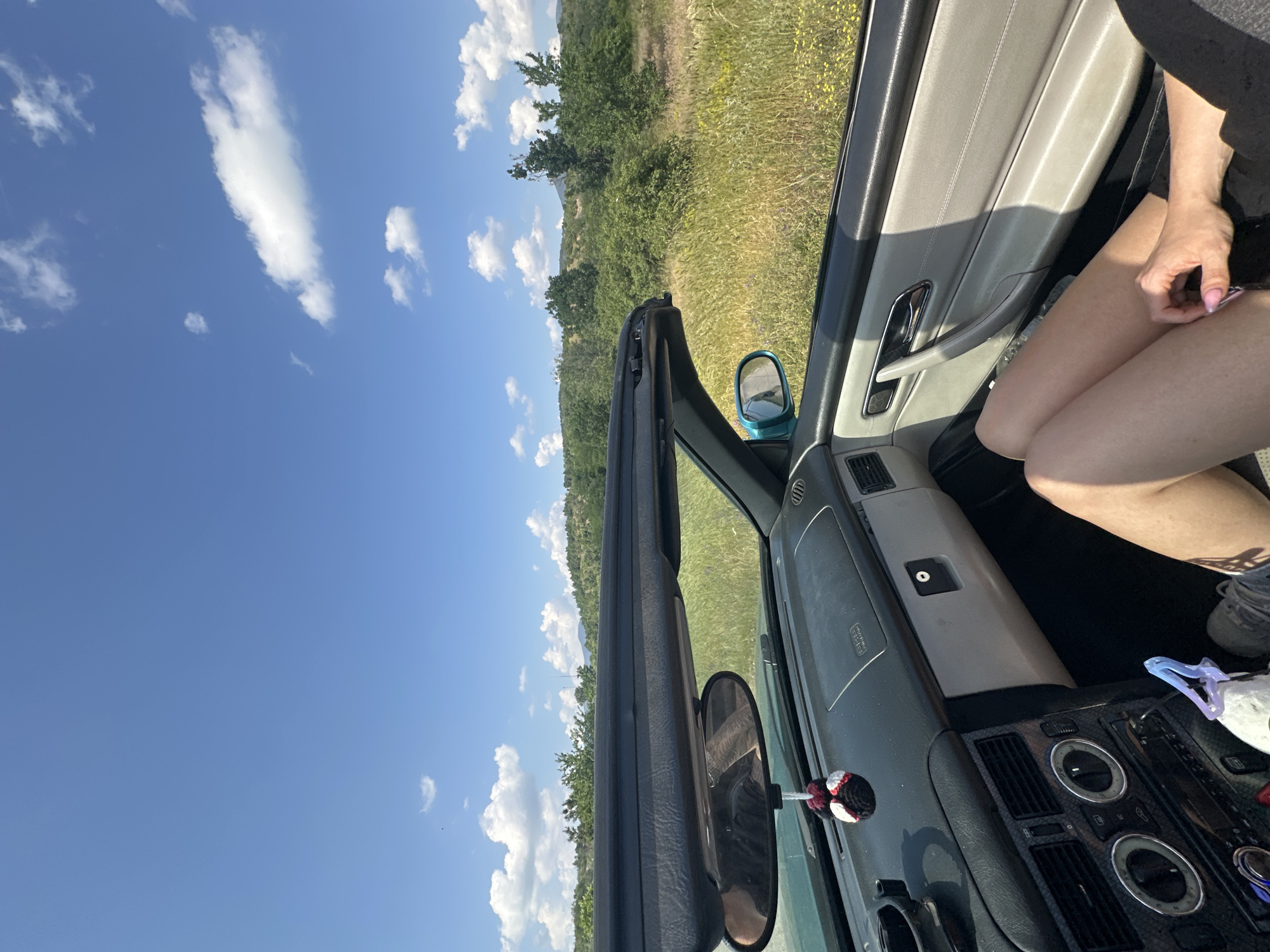 The image captures a first-person perspective from inside a convertible car on a bright, sunny day, with the scene heavily rotated counter-clockwise. The car's interior details are visible, including a dark dashboard with circular controls and air vents, a silver door handle on a lighter grey door panel, and a side mirror. A small, black and red, spider-like plushie hangs from the rearview mirror. A person's bare legs, partially covered by shorts, and one hand are visible in the foreground on the right, suggesting they are a passenger or driver enjoying the ride.
Outside the car, a vibrant blue sky dominates the upper portion of the frame (when corrected for rotation), adorned with scattered white, fluffy clouds. The landscape unfolds as a series of green hills covered in lush vegetation, including dense trees and fields of tall grass dotted with yellow wildflowers. The setting suggests a rural or semi-rural area, consistent with the Cherganovo region of Bulgaria. The clear sky and bright sunlight indicate pleasant weather, ideal for a convertible drive. No specific text is clearly legible within the image.