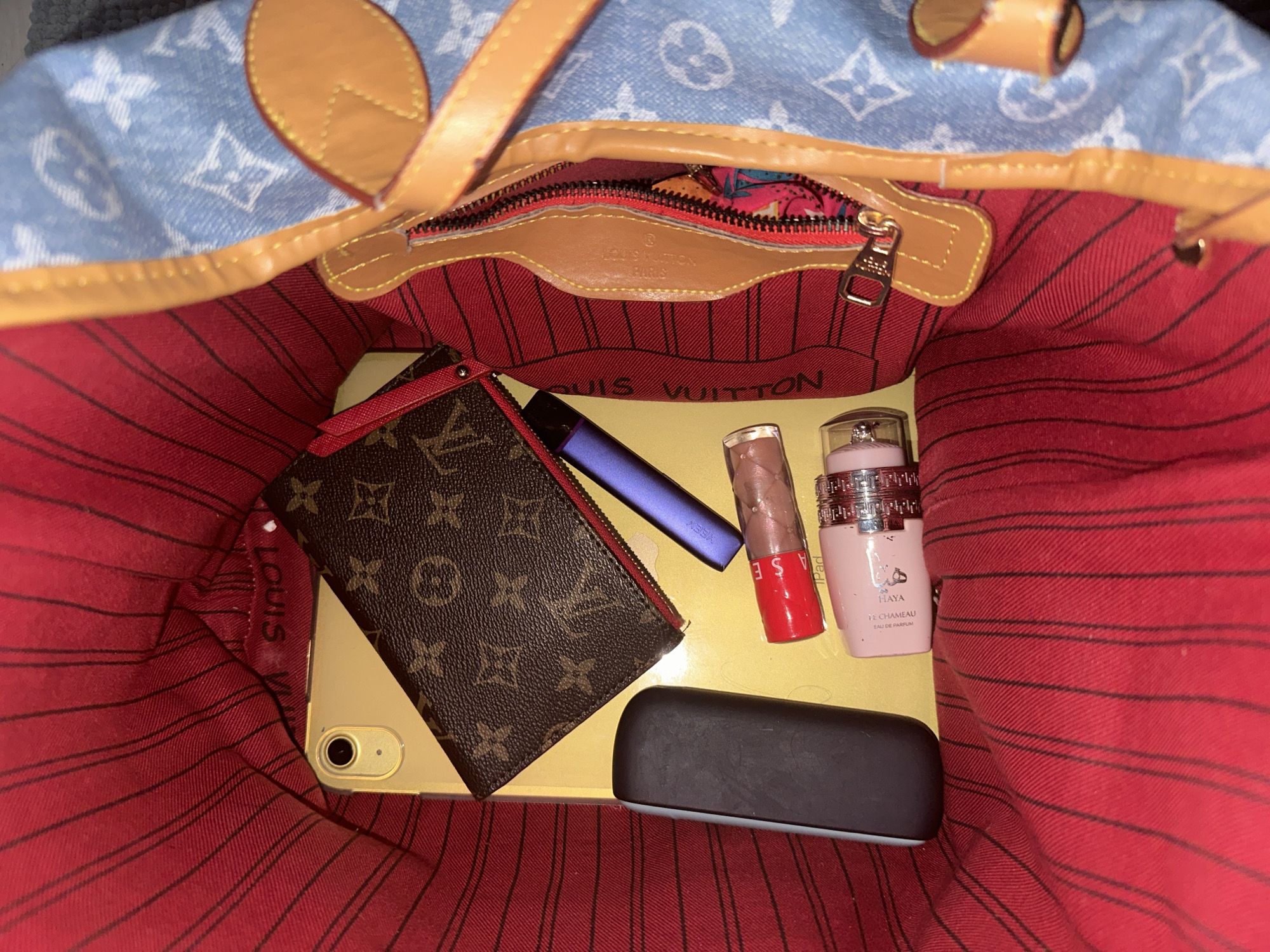 The image shows the interior of a Louis Vuitton Neverfull MM handbag. Inside the bag, on top of what appears to be an iPad, there is a Louis Vuitton wallet, a vape pen, a lip gloss, a perfume bottle labeled "Haya Le Chameau," and a sunglasses case. The interior of the handbag is red with dark brown stripes, and the exterior is light blue with white Louis Vuitton logos. The photo does not provide information about the location.