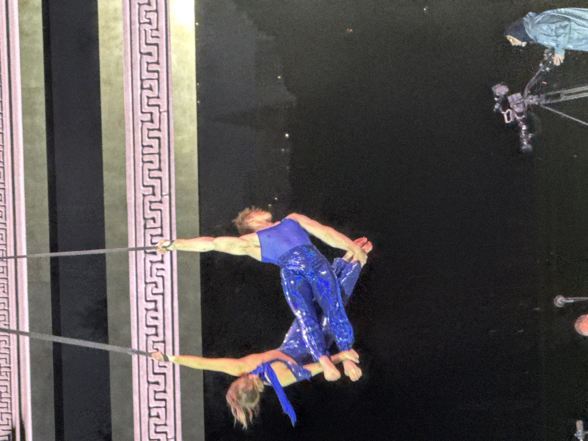 Two aerial acrobats, one male and one female, perform on aerial silks in front of a dark background. The male acrobat is on the right, wearing a blue sequined jumpsuit. The female acrobat is on the left, also in a blue sequined outfit, with a blue sash. Both performers are suspended by ropes. In the lower right portion of the image, a person in a hooded jacket operates a video camera on a tripod. Microphones are also visible in the foreground. The background features horizontal bands of patterned decoration. The scene appears to be an outdoor performance or event.