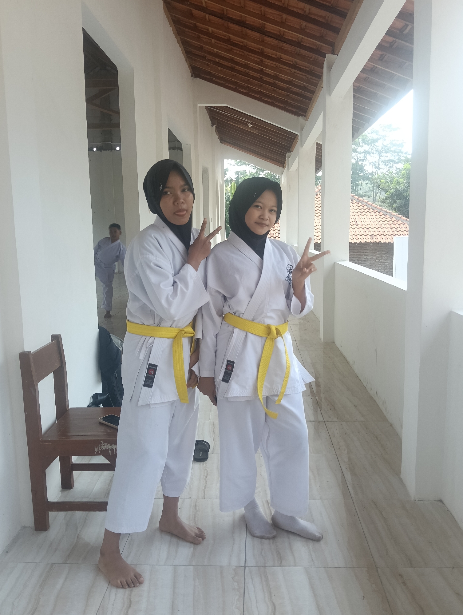 Two young women wearing white karate uniforms with yellow belts stand in a covered outdoor walkway in Adinuso, Indonesia. Both are wearing black hijabs and are posing with peace signs. The woman on the left is sticking out her tongue. The walkway has white pillars and a wooden roof. In the background, another person in a white karate uniform can be seen walking away, and a tiled roof building is visible beyond the walkway. A wooden chair is positioned to the left of the women.