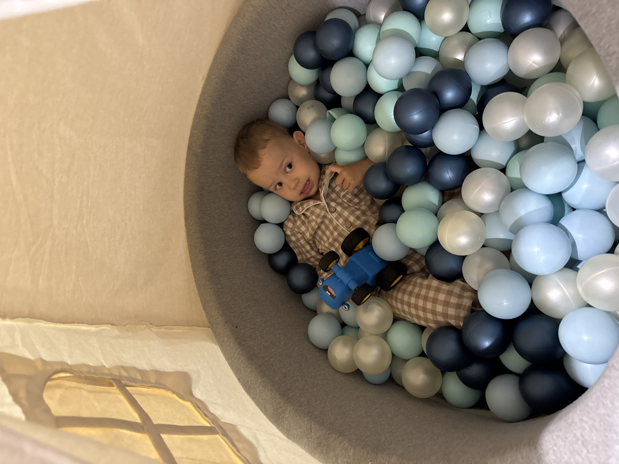 A toddler in a brown and white checkered pajama shirt is lying in a ball pit filled with blue, light blue, and white balls, while holding a blue toy truck. The pit is gray and appears to be part of a play tent. The scene is inside, with a window visible at the bottom left. The specific location in the city of Rusanovo, Russia, cannot be determined from the image.