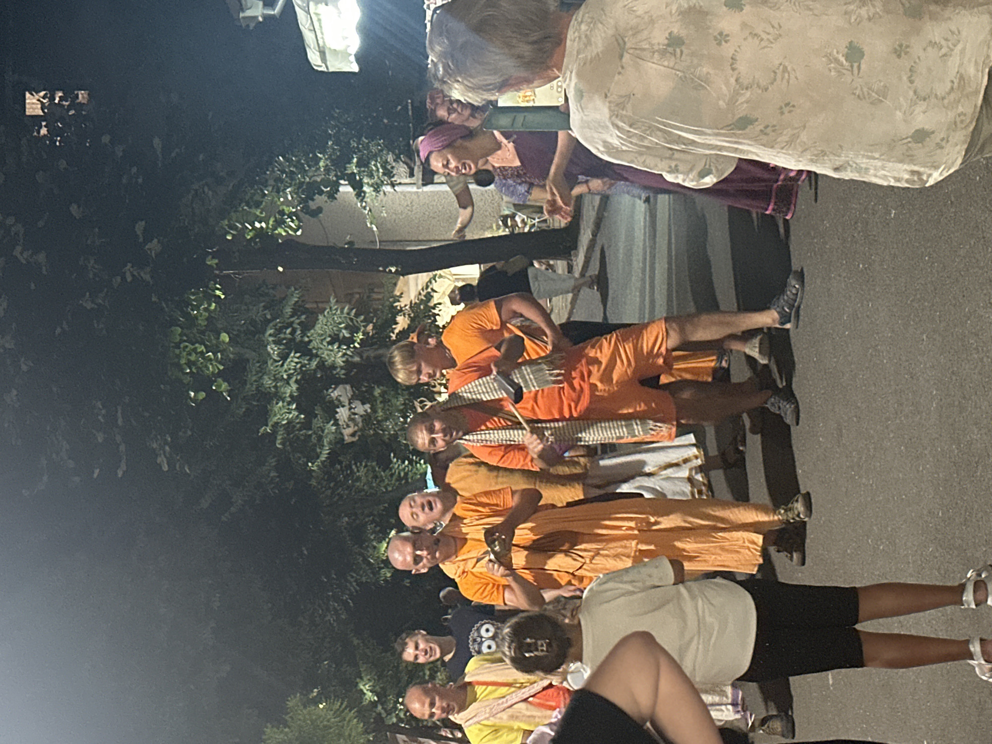 The image shows a group of people gathered on a street at night in Tsarevo, Bulgaria, likely for a religious or cultural event. Several men in orange robes are singing or chanting, with one holding a small metal instrument. Another man is holding a drumstick. A young girl in a white shirt and black shorts stands in the foreground. To the right, a woman in a purple dress and headscarf is speaking or singing, with her hands gesturing. The background shows trees and buildings, with some street lighting. The ground is paved asphalt.