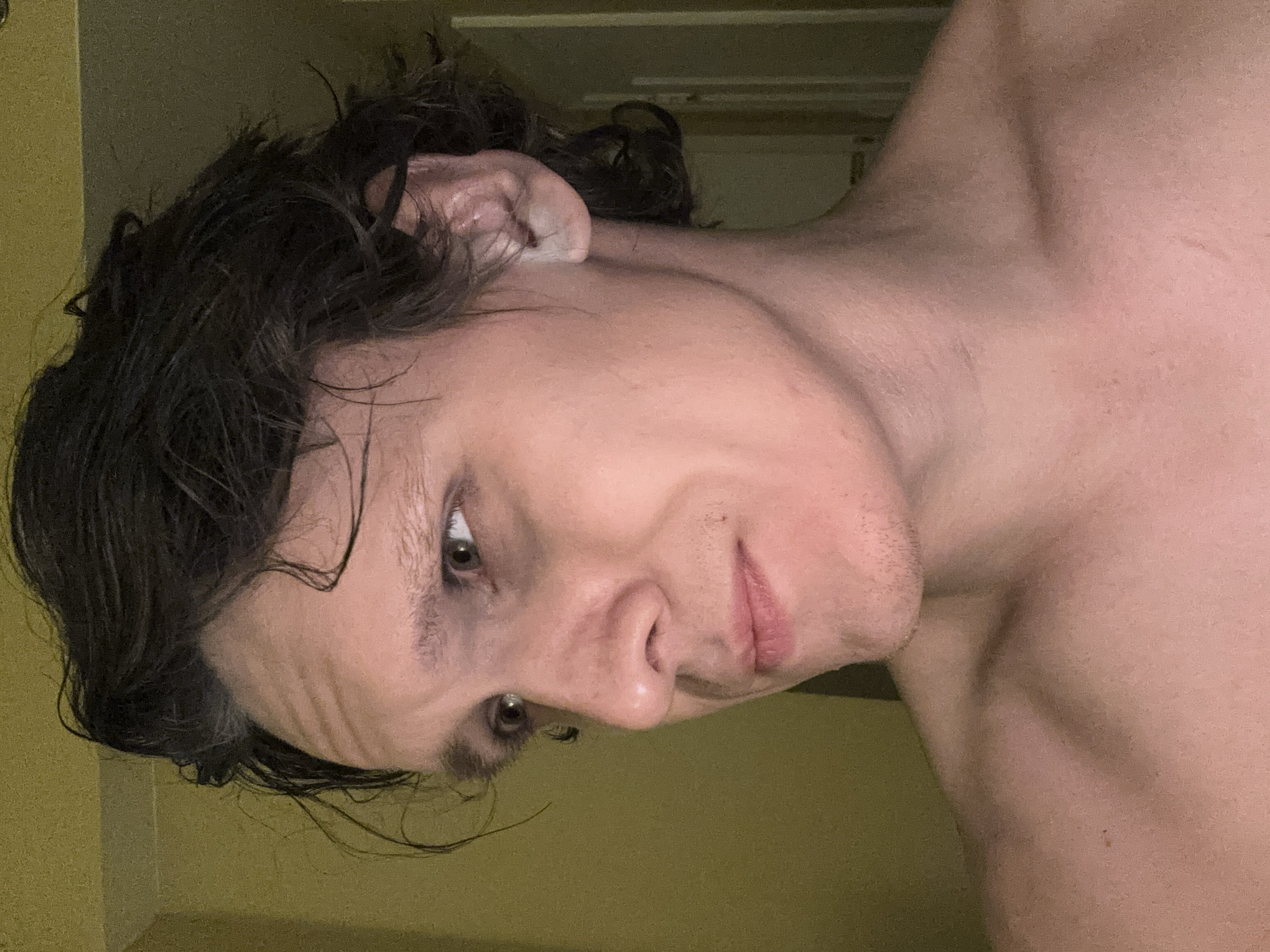 A close-up, profile shot of a light-skinned individual with dark, unkempt hair is captured from the chest up. The subject is looking to the left, with their head tilted slightly down and to the side. Their eyes are open and looking away from the camera. The lighting in the image is dim and warm, with shadows cast on the subject's face. The background appears to be a wall with a textured, yellowish hue and a doorway in the distance. The individual's shoulders and upper chest are visible and bare. There are no discernible objects or activities taking place in the scene. The image does not provide any specific information to determine its location in San Francisco, United States of America.