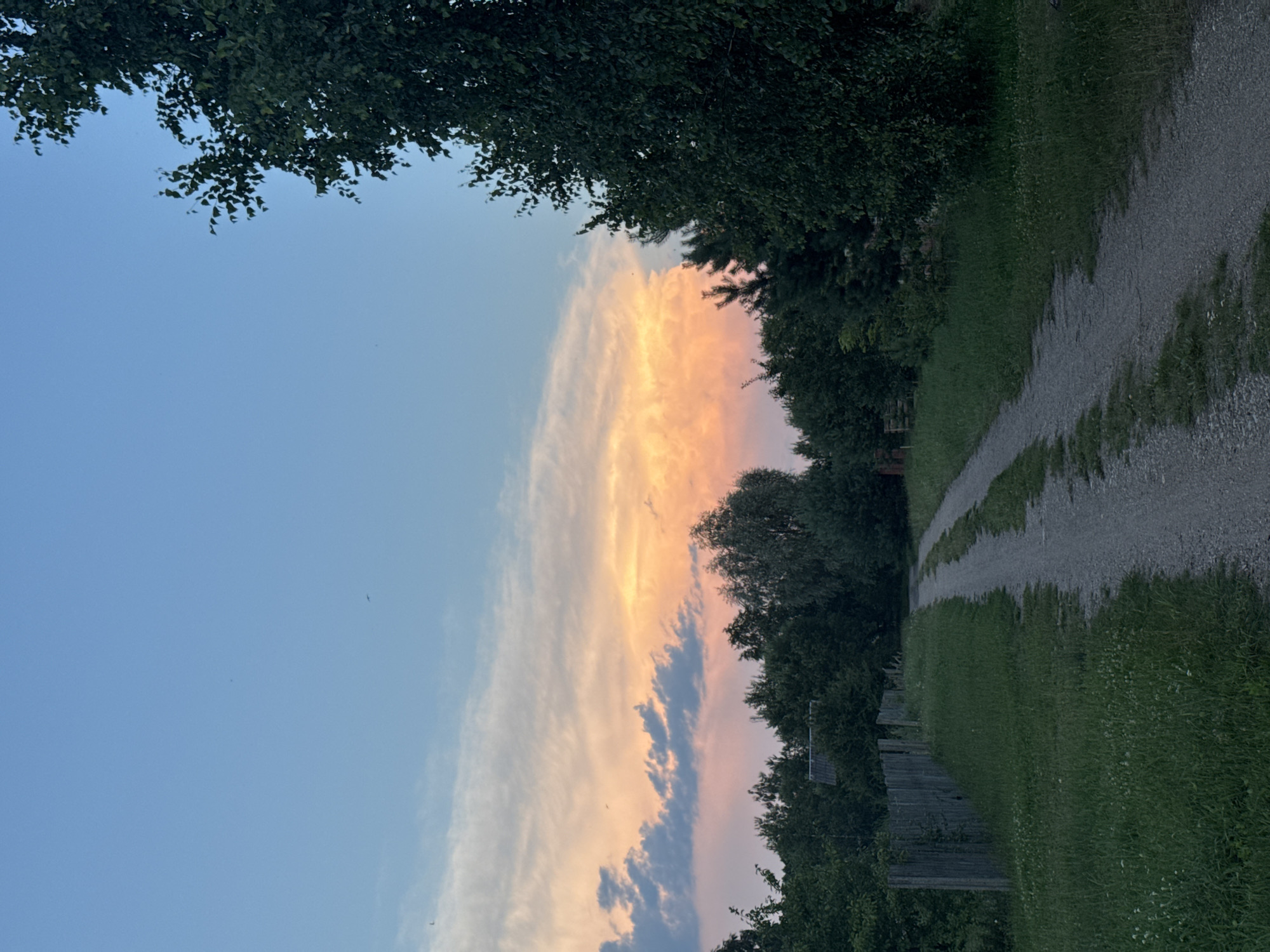 This media file depicts a rural landscape in the Neman rural council, Belarus, during what appears to be sunset. The scene is dominated by a path made of gravel and grass, winding through lush greenery and bordered by a rustic wooden fence on one side. Dense foliage from trees frames the left and right edges of the image.
The sky is a prominent feature, displaying a gradient of colors from a clear, pale blue on the left to vibrant shades of orange and pink in a band of clouds stretching across the center. Darker clouds are interspersed within this colorful display, adding depth and texture to the sky. The lighting suggests the late evening or early morning, with the soft glow of the setting or rising sun illuminating the clouds.
The overall mood of the image is tranquil and serene, evoking a sense of peaceful rural life. The absence of any visible text or human figures further enhances this quiet atmosphere. The natural elements – the path, grass, trees, and sky – are the sole subjects, creating a simple yet captivating natural scene.